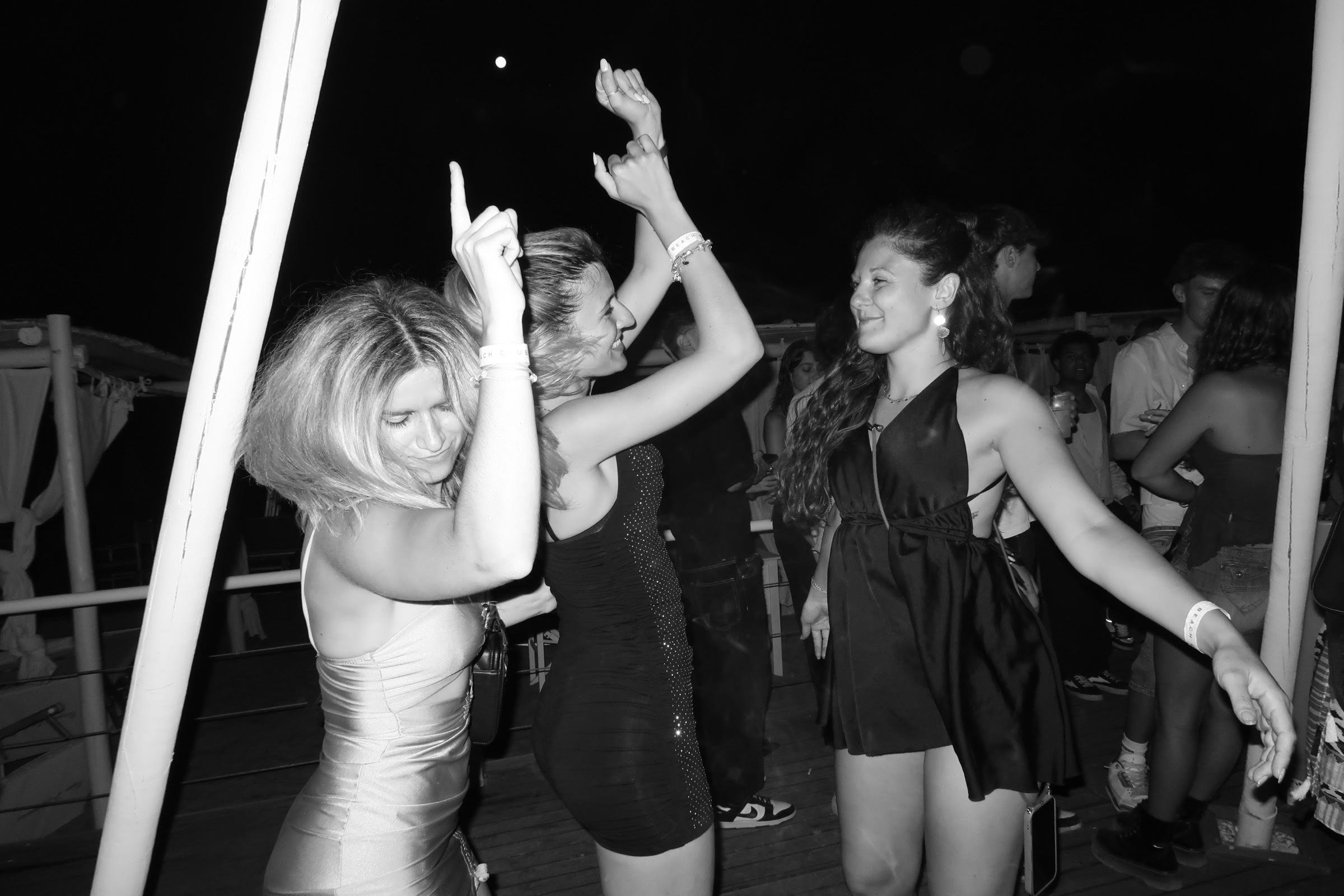Four women dancing and having fun at a party or nightclub at night, with a dark background and people in the background.