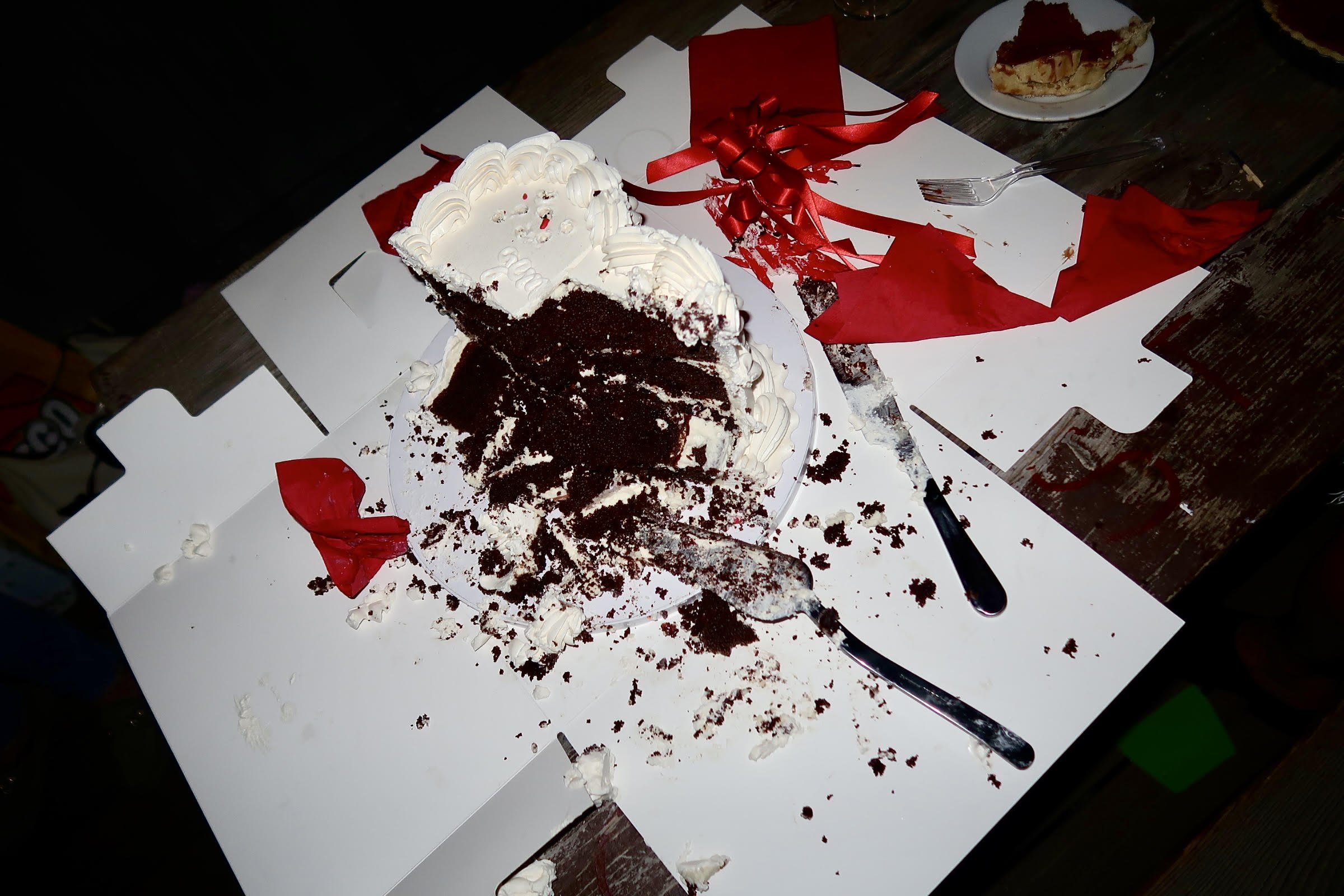 A partially eaten chocolate and cream layered cake on a platter, surrounded by crumbs and remnants of red and white ribbons on a cluttered table with a fork, a knife, and a plate with a slice of pie.