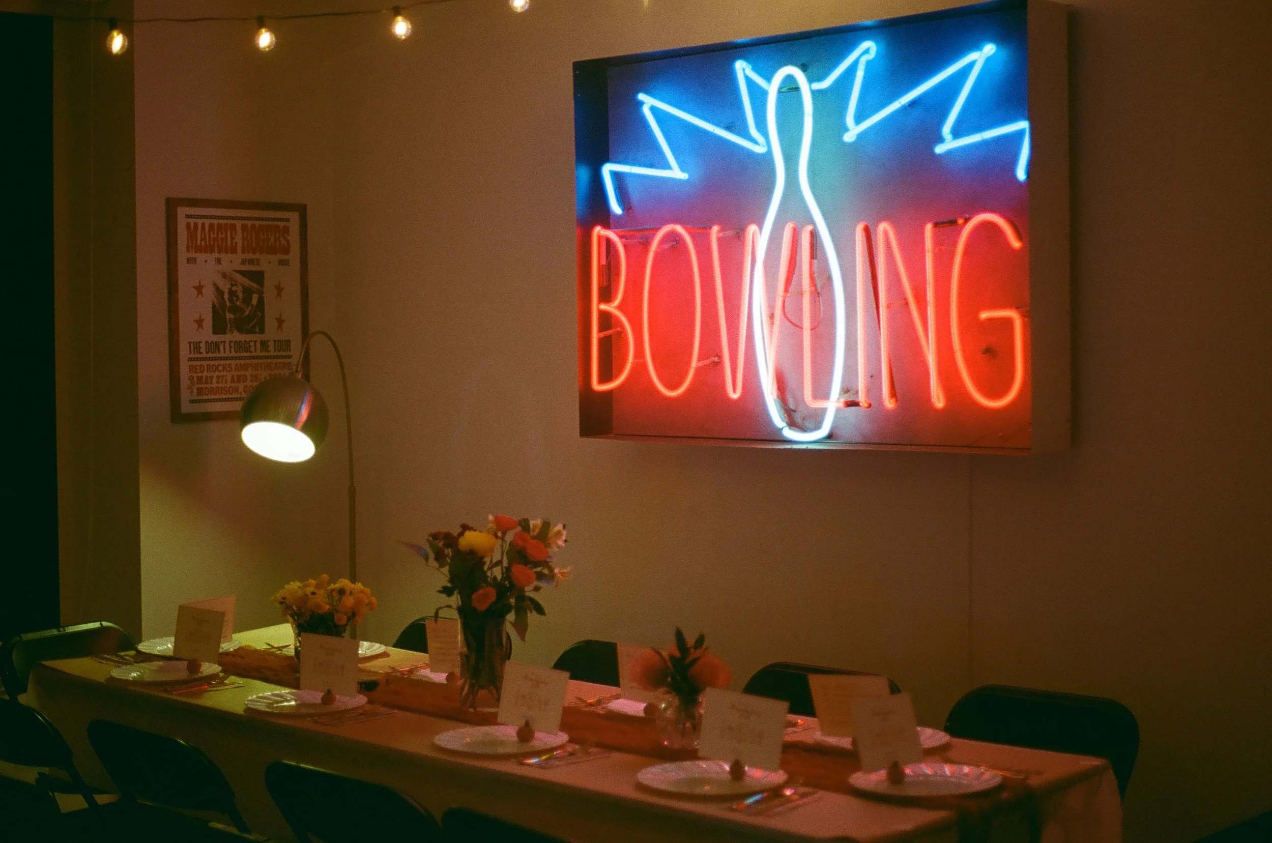 A dining setup with a long table decorated with flowers and place cards, illuminated by warm lighting. There is a neon sign on the wall that says 'BONING' with a graphic of a bird above it. The background features a poster on the wall and a table lam