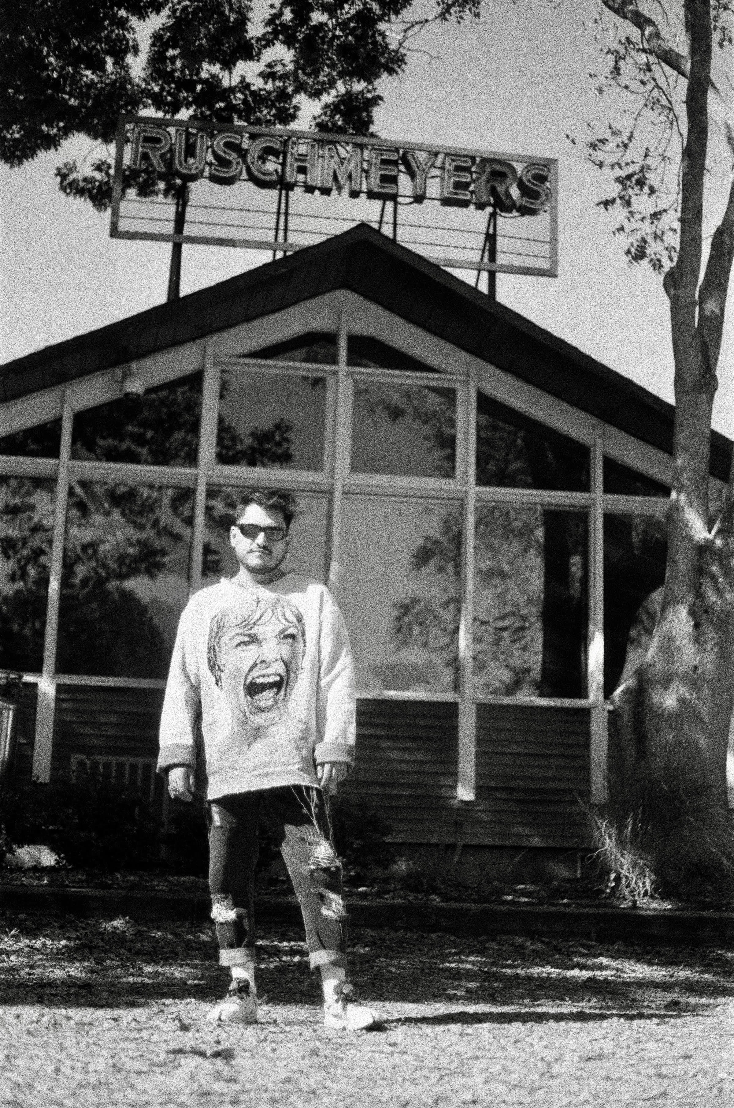 A young man with sunglasses wearing a long-sleeved sweatshirt with a woman's face with an open-mouthed smile printed on it. He is standing outdoors on a dirt path with trees around, and a house with a sign that reads 'RUSCHMEYERS' on the roof in the 