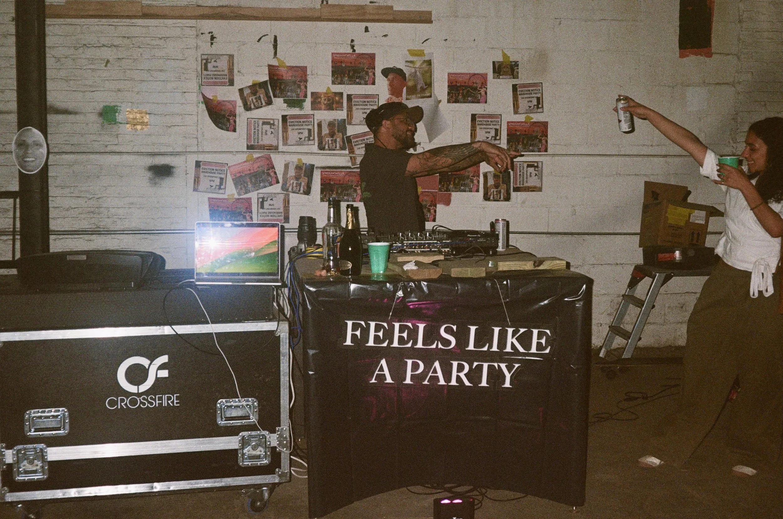 Two people are dancing and drinking at a party. The man in the center with tattoos and a cap is DJing with equipment on a table, while the woman on the right holds a beer can and reaches out. The background has a wall decorated with flyers and poster