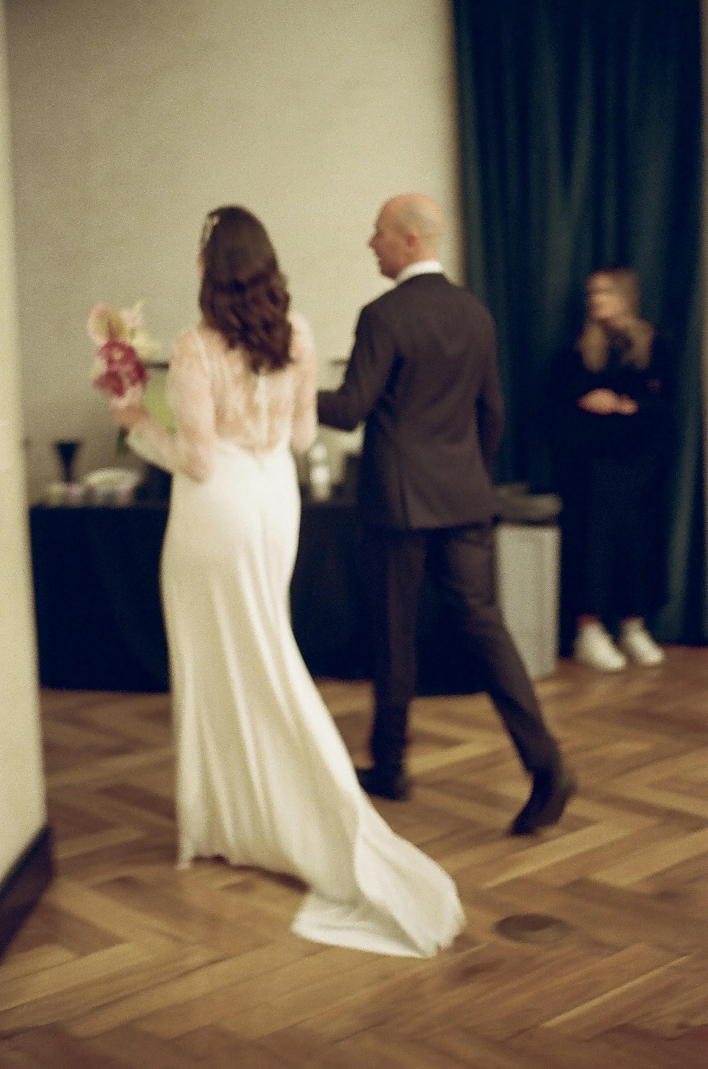 A bride in a white gown holding a bouquet faces a groom in a black suit. They appear to be exchanging vows or presenting at a wedding ceremony in an indoor setting with dark curtains and wooden flooring. In the background, a person is sitting on a chair.