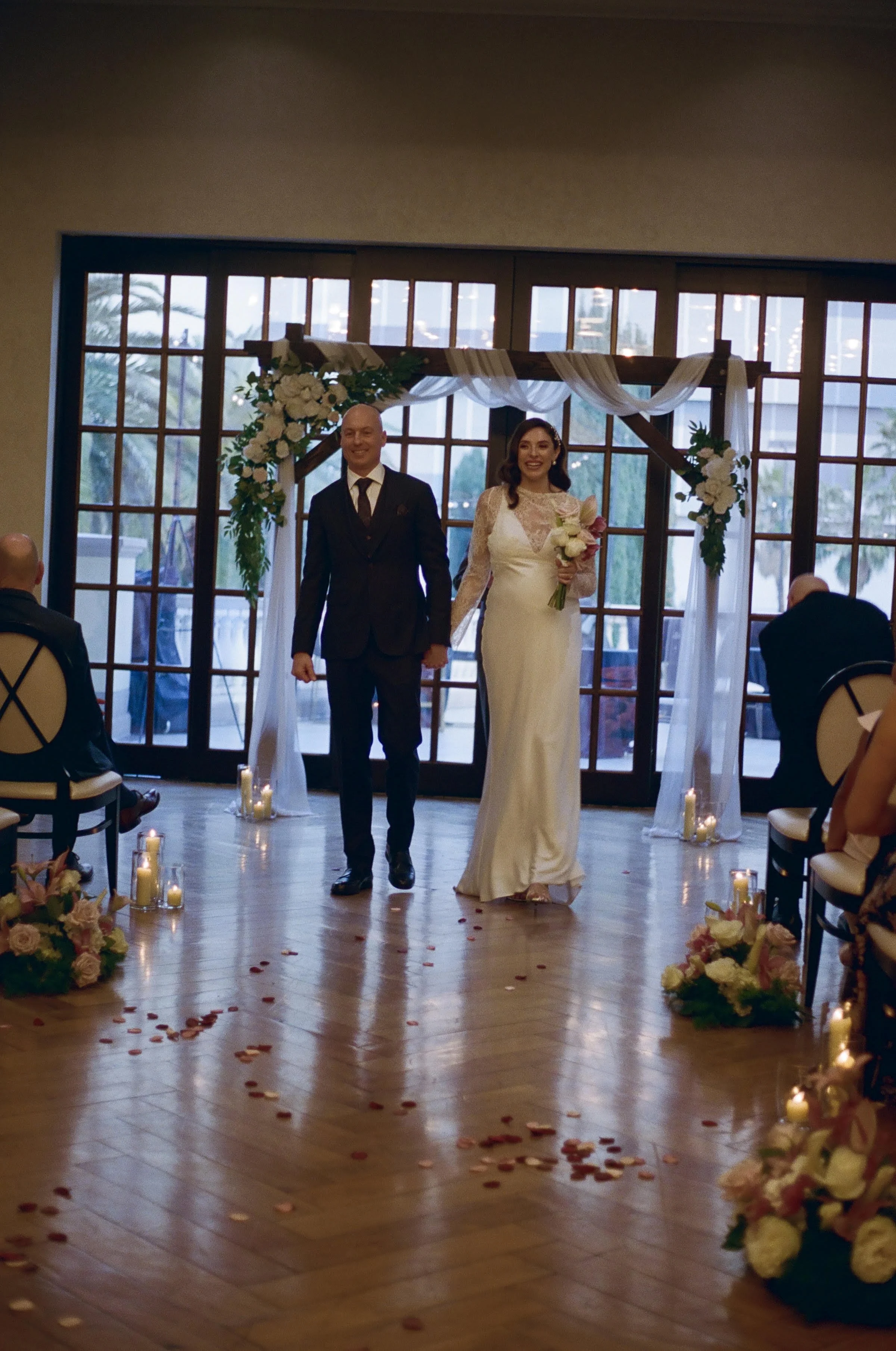 Bride and groom standing hand in hand in a wedding venue with wooden floors, candles, and flower arrangements, smiling, after exchanging vows.