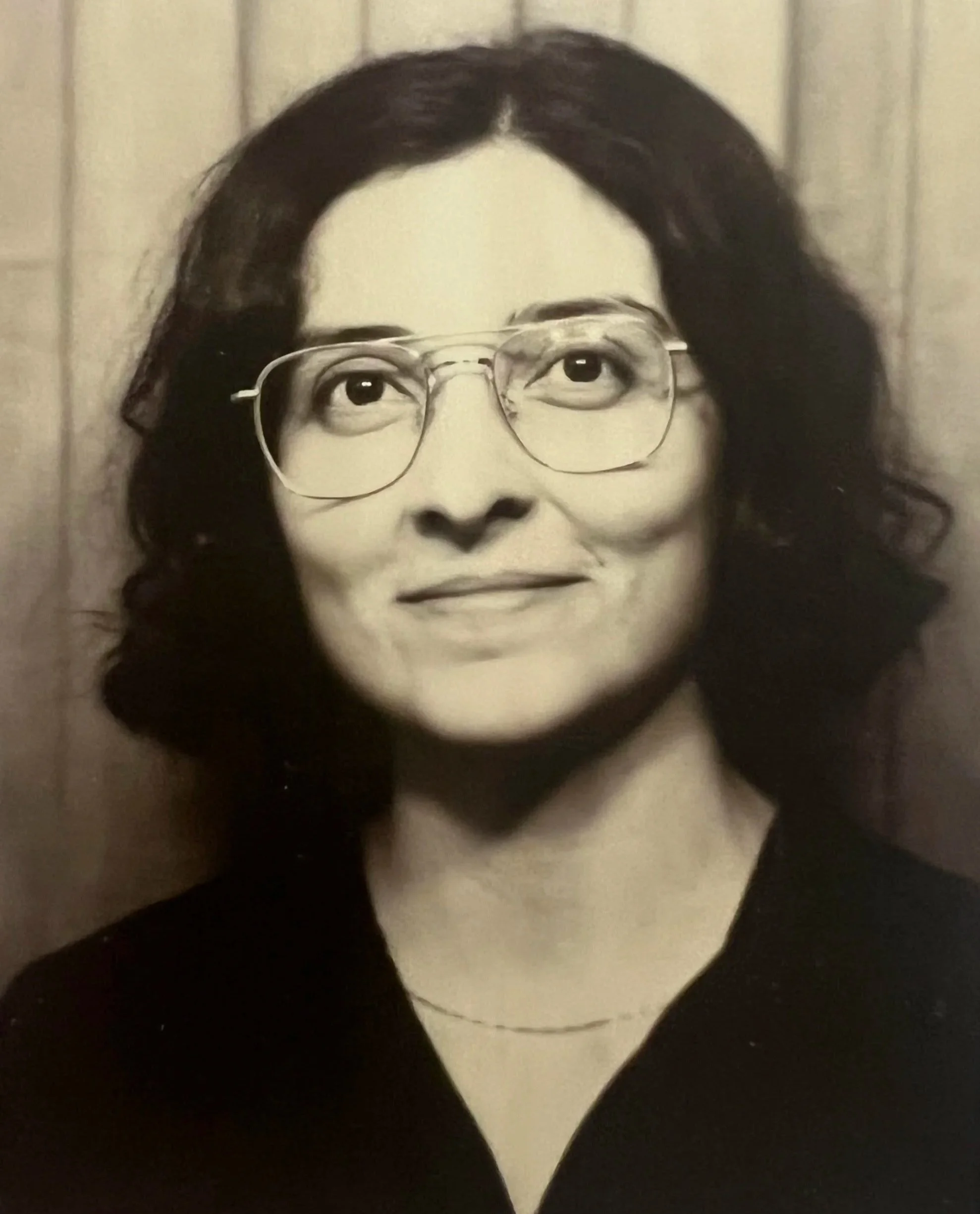 A black-and-white photograph of a woman with dark curly hair and glasses, smiling, against a background of wooden panels.