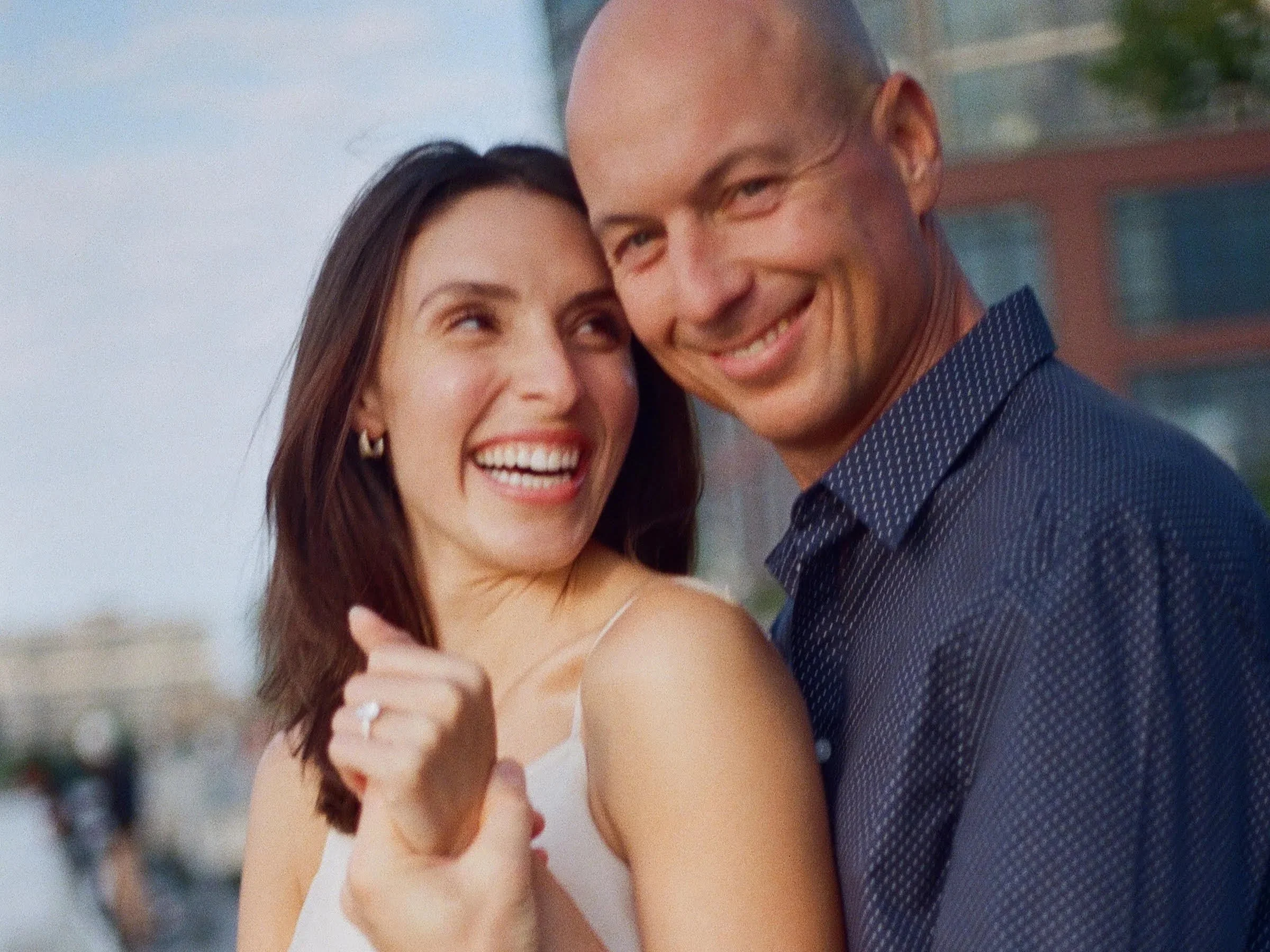 A happy couple, a woman with dark brown hair and a man with a bald head, smiling and embracing each other outdoors on a sunny day.