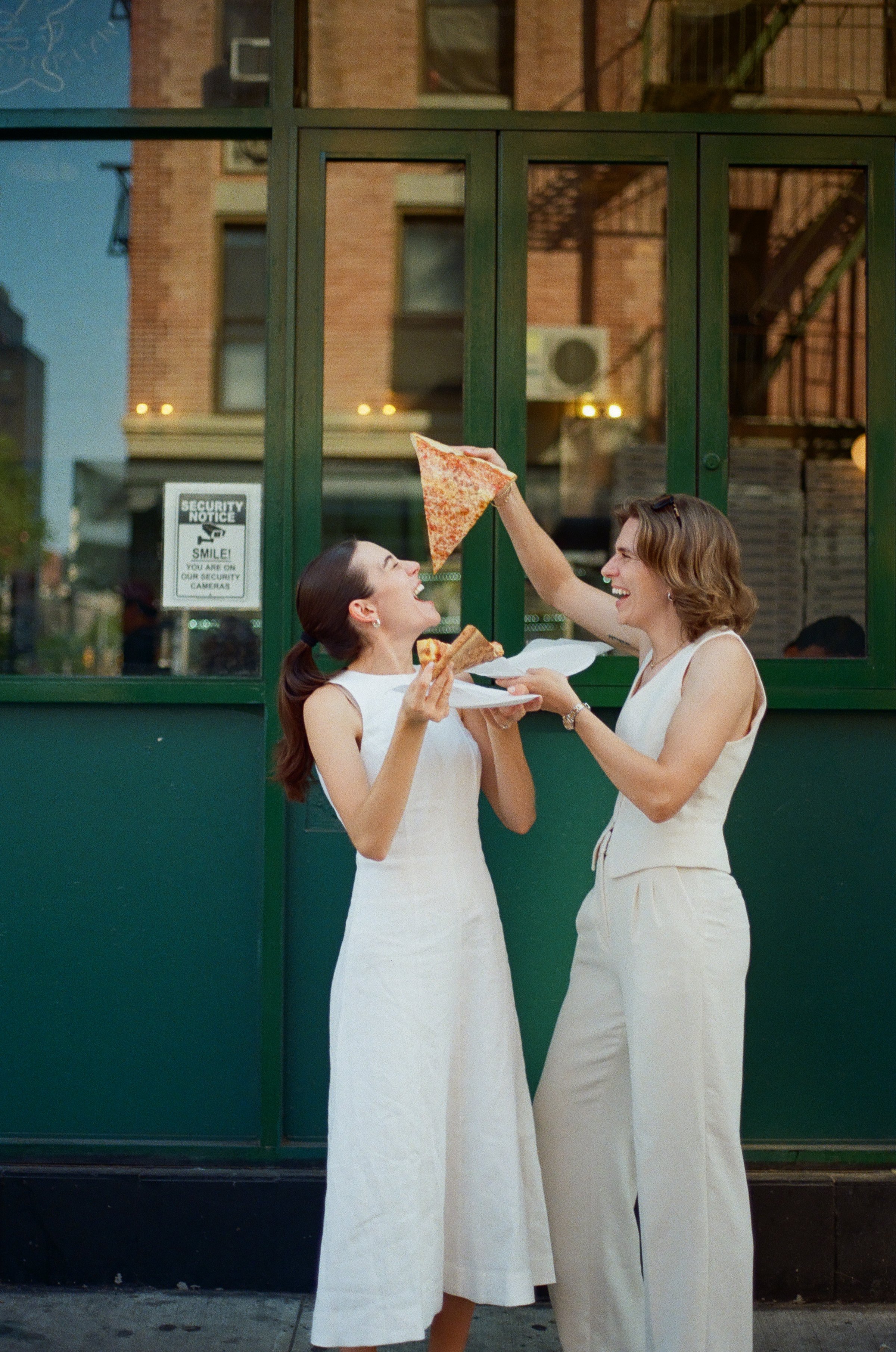 Two women wearing white dresses sharing a slice of pizza, standing in front of a green window with a city building in the background.