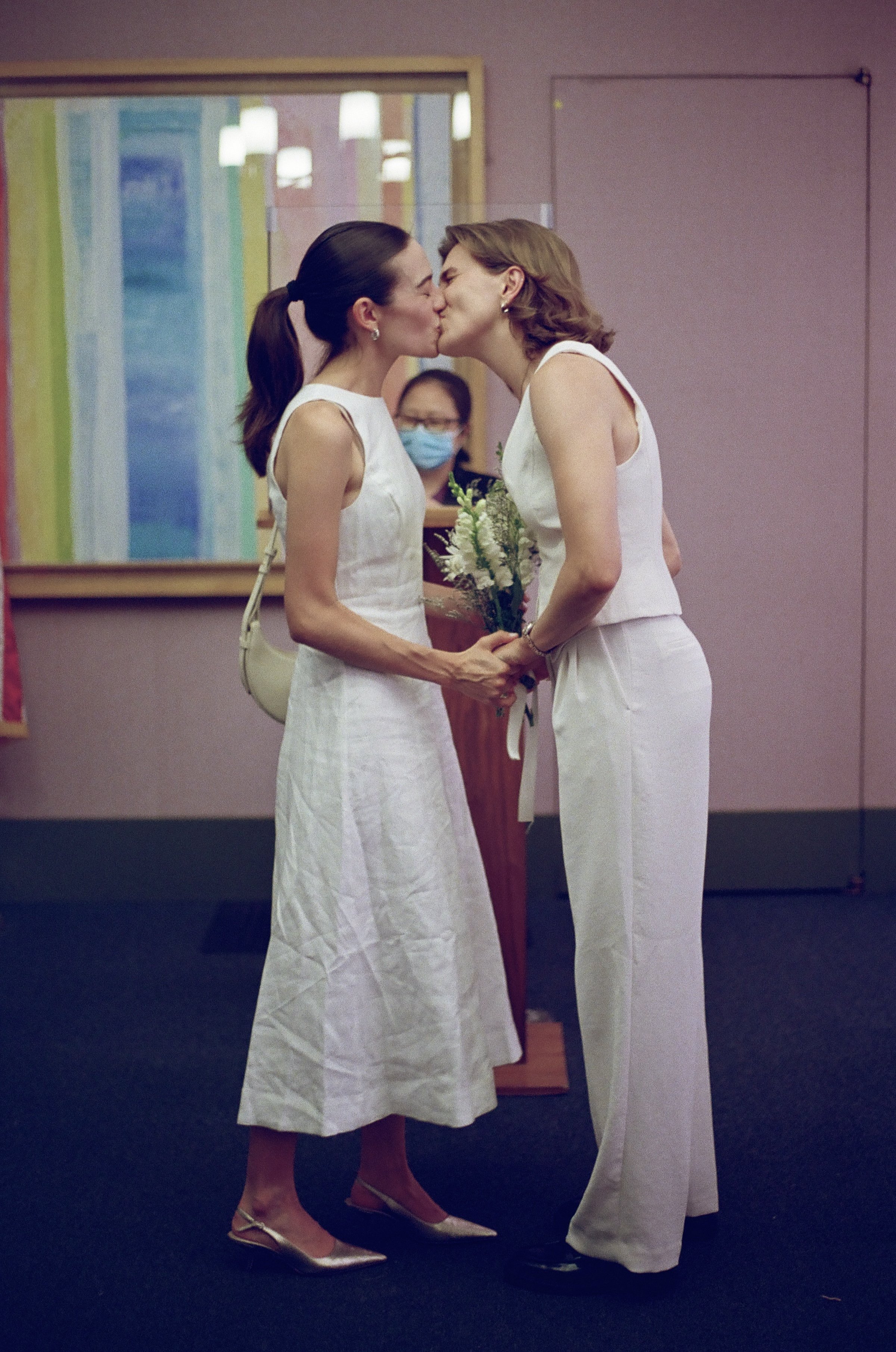 Two women in white dresses share a kiss while holding hands, with a woman wearing a face mask standing in the background.