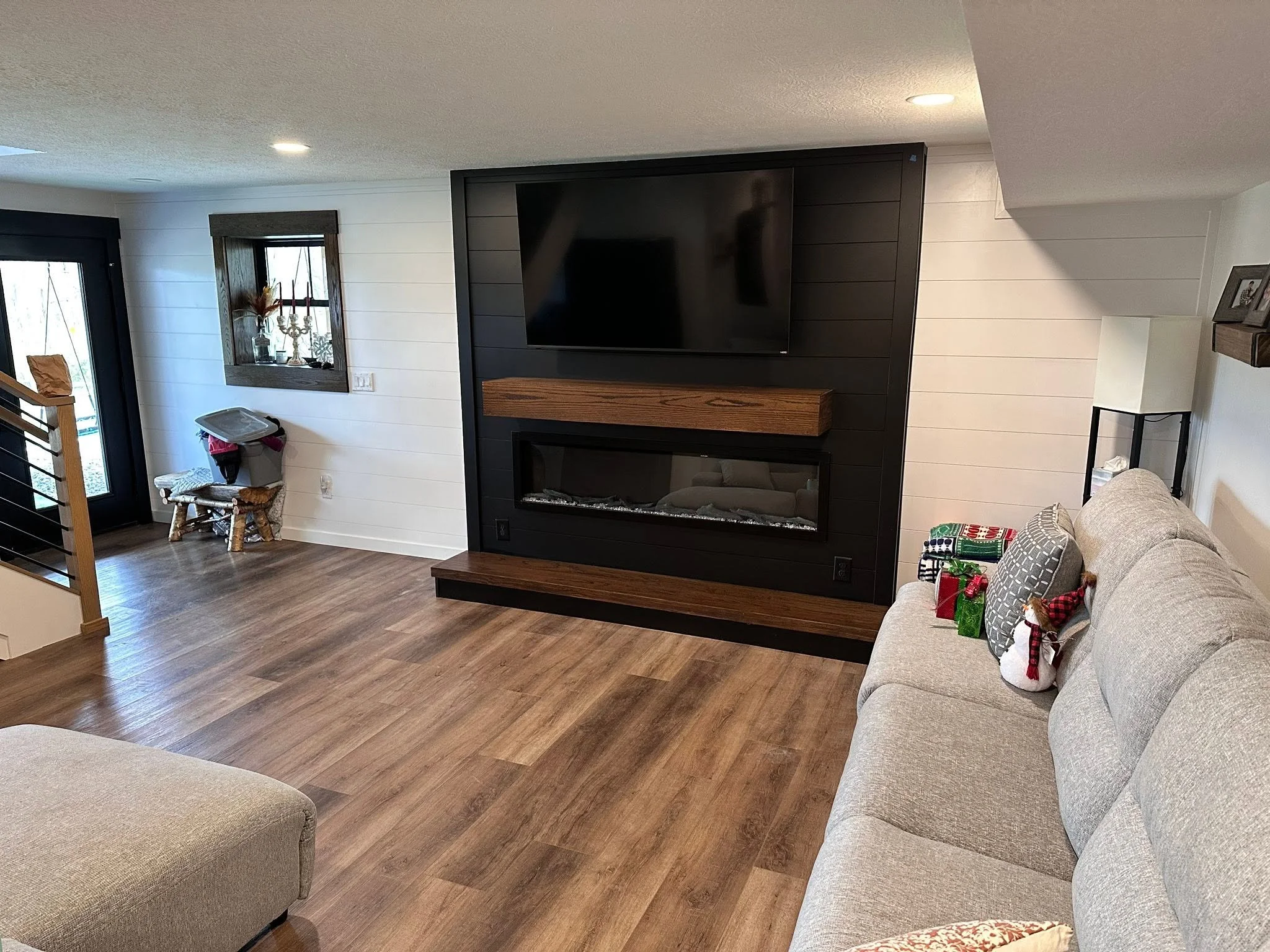 A living room with wooden flooring, a gray couch with holiday-themed pillows, a firepit in a black frame with a wooden mantel, a wall-mounted flat-screen TV, and decorative items on small shelves and window ledges.