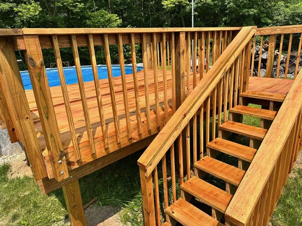 Wooden deck with steps, overlooking a backyard with grass, trees, and an above-ground swimming pool with a blue liner.