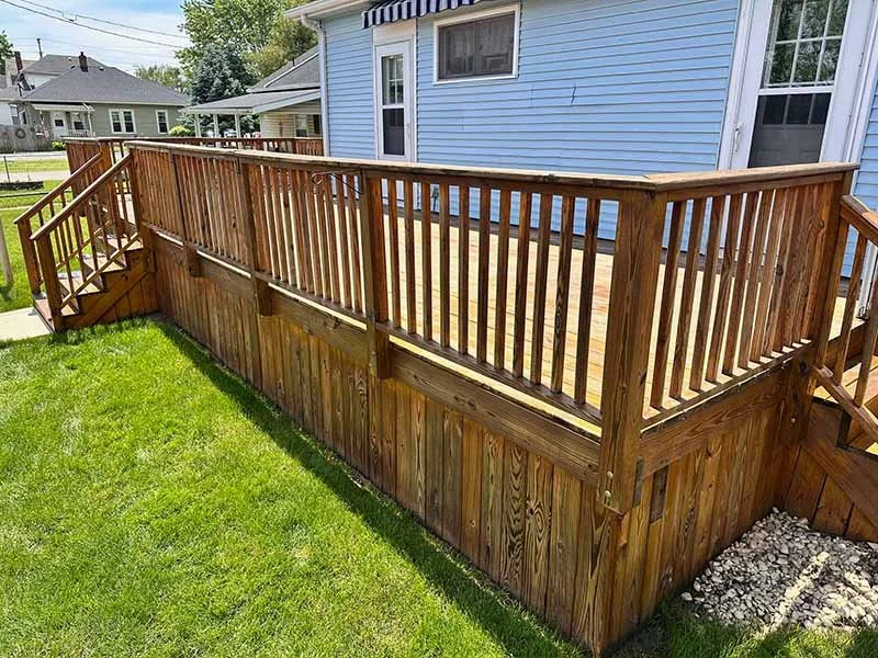 Wooden deck with railing attached to a blue house, stairs leading down to a grassy yard, and neighboring houses in the background.