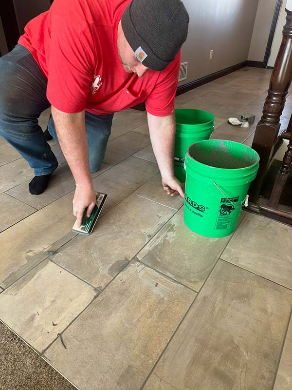 A worker using a trowel to install new large beige tiles. There are two green buckets nearby filled with grout or adhesive, indicating a tile installation project.
