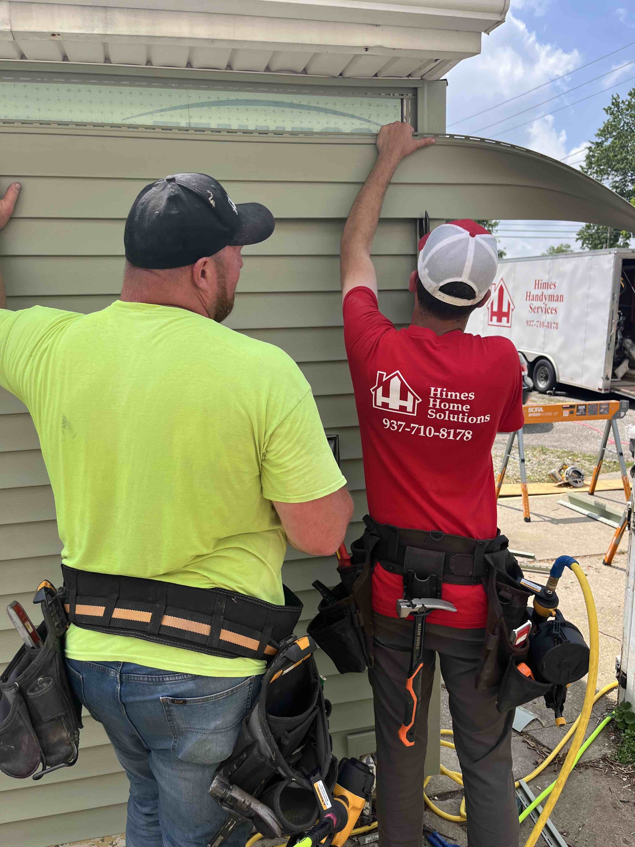 Two workers are installing or repairing the exterior siding of a house. They are working together, using tools near a window.