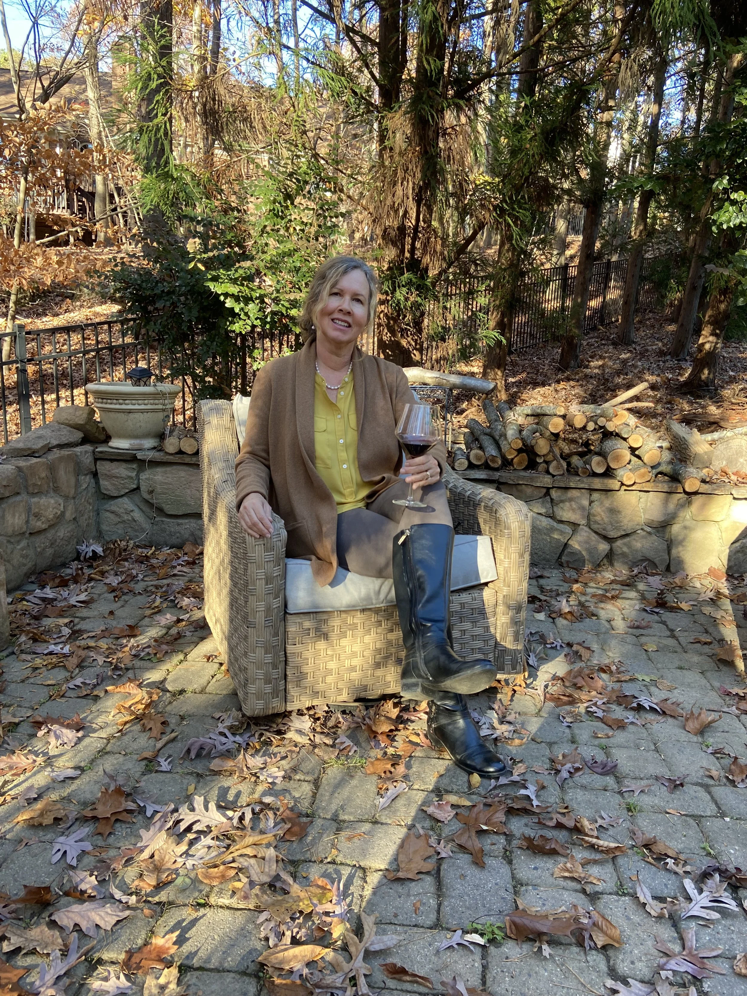A woman sitting outdoors on a wicker chair, holding a glass of red wine, surrounded by fallen autumn leaves and trees with a fence in the background.