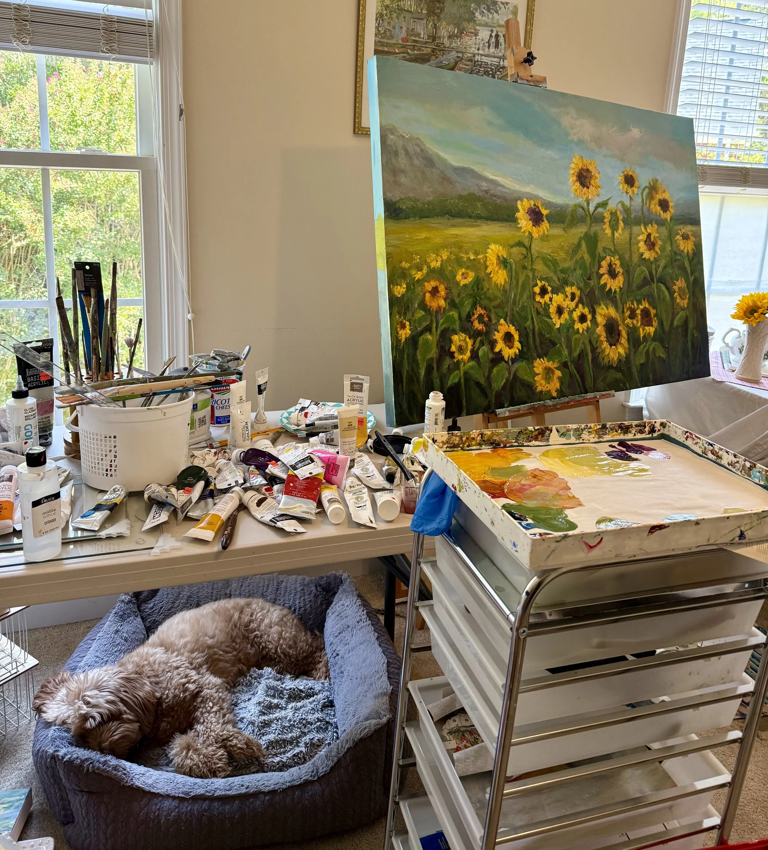 An indoor art studio with a large landscape oil painting of a field of sunflowers with mountains and cloudy sky behind it. The studio has two windows with blinds, a cluttered table with art supplies, and a plush dog bed with a large brown and white poodle lying on it. An easel stands next to the painting, and there are various painting tools and tubes of acrylic paint scattered around.