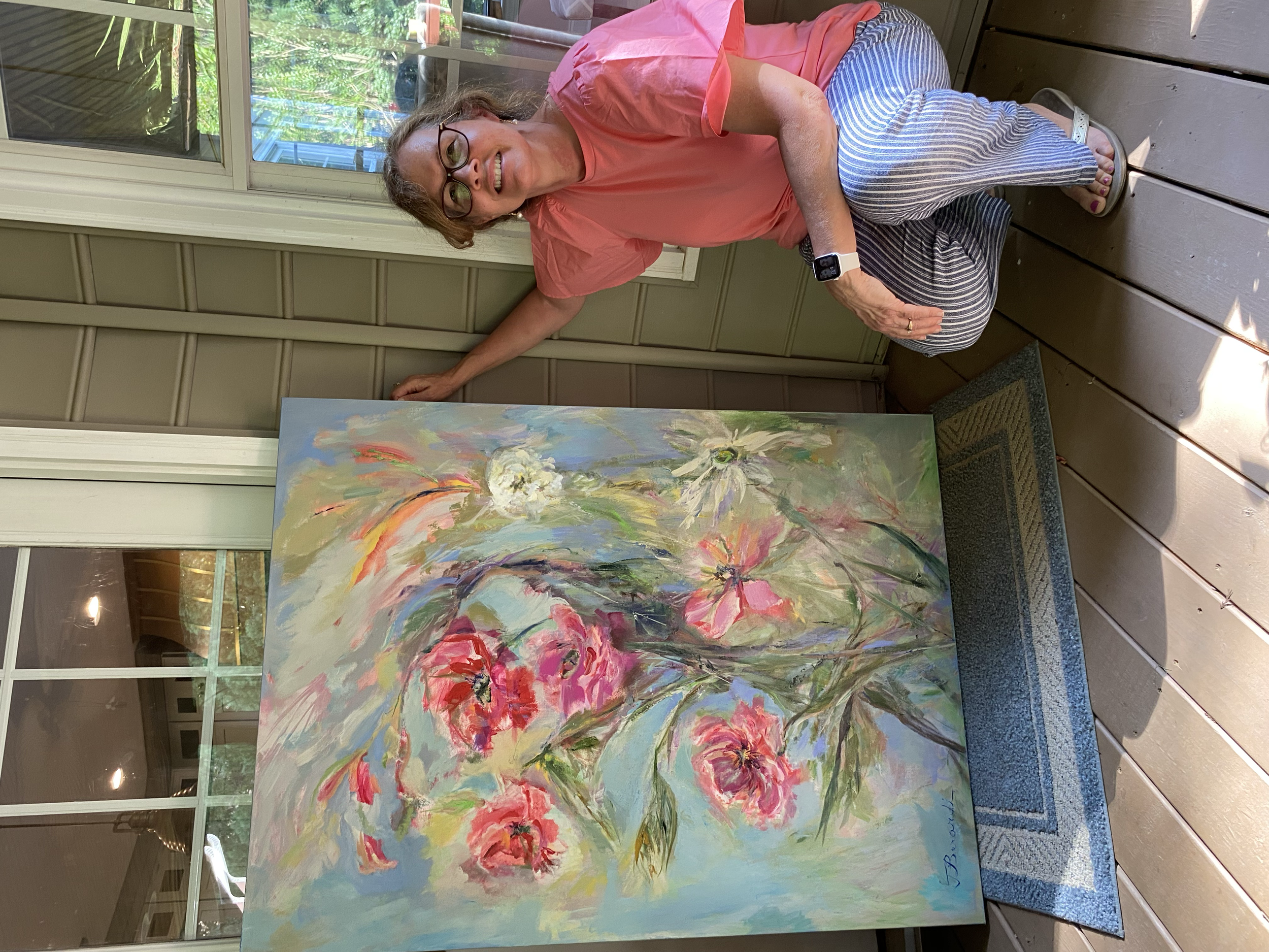 A woman in a pink shirt, striped pants, and sandals is kneeling next to an abstract floral painting and smiling at the camera. The painting features large pink, white, and purple flowers with green leaves on a blue background. The setting appears to be indoors near a window.