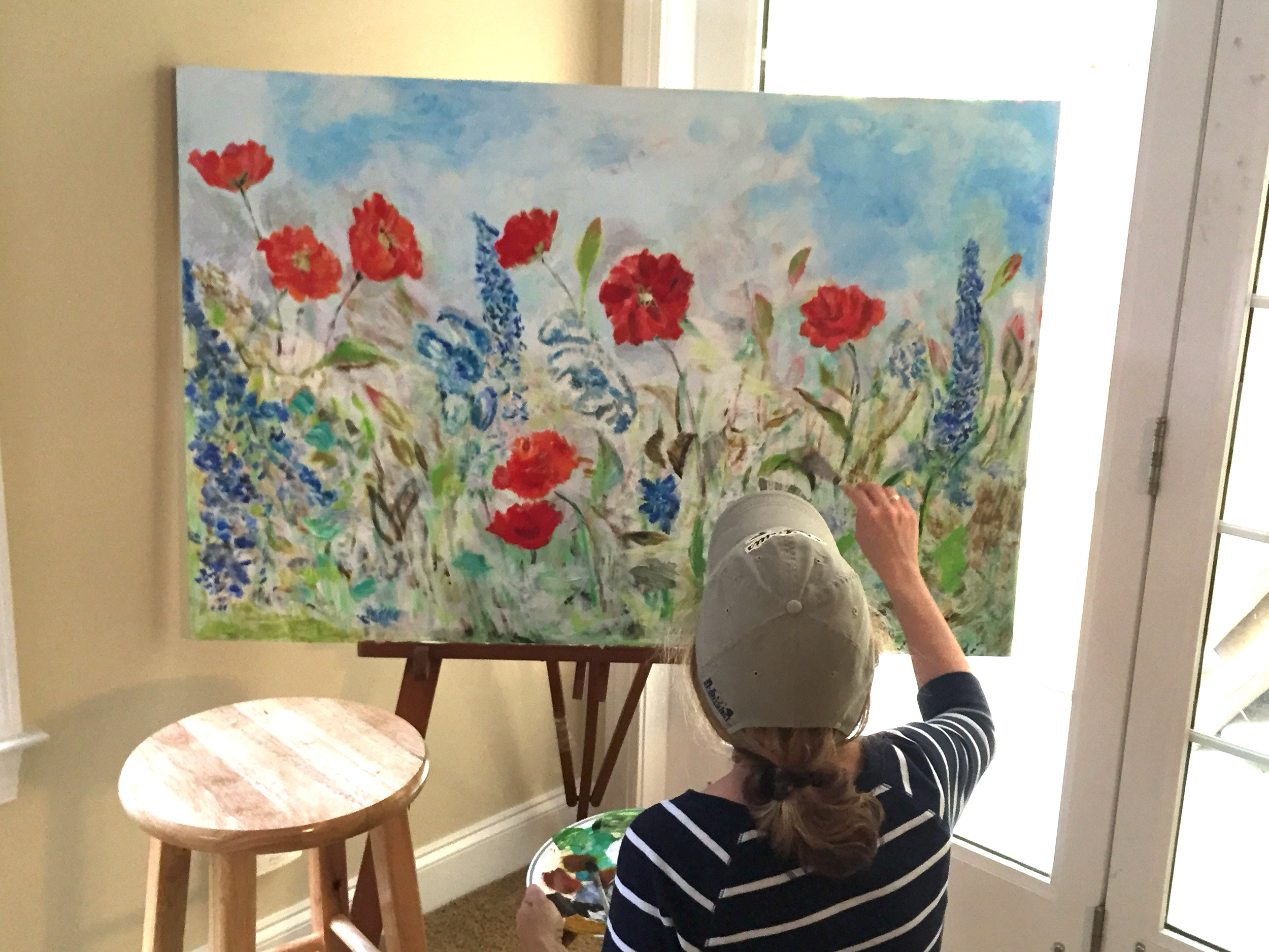 A woman painting a large canvas with flowers in an indoor setting. The painting features red flowers and greenery against a sky background. The woman wears a striped shirt and a cap, holding a palette.