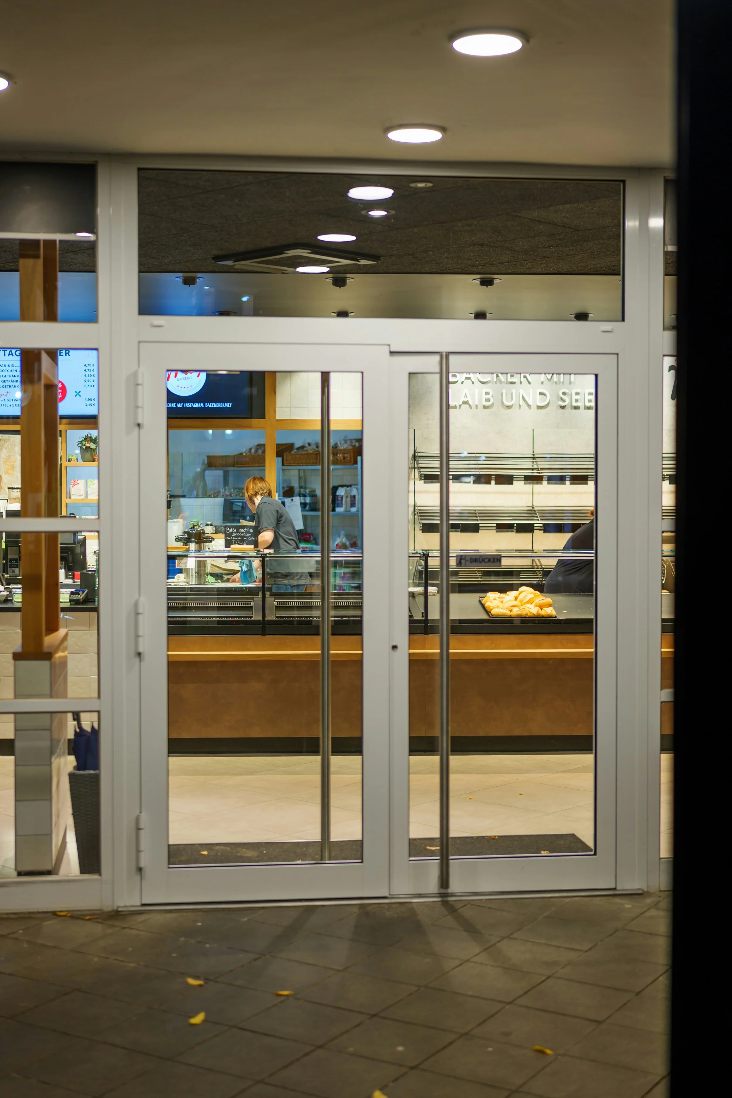 Entrance to a store, viewed through glass doors, with a person inside preparing food and bread on a tray on the counter.