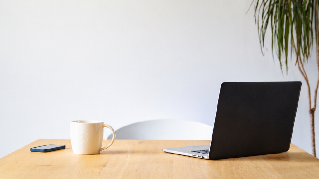 A wooden table with a closed silver laptop, a black smartphone, and a white mug, with a white chair and a green plant in the background.