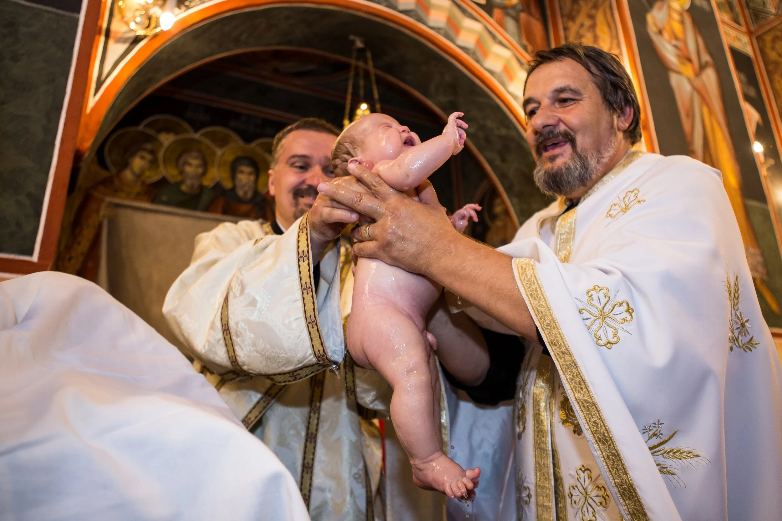 A priest and another man holding a baby during a baptism ceremony in an ornately decorated church.