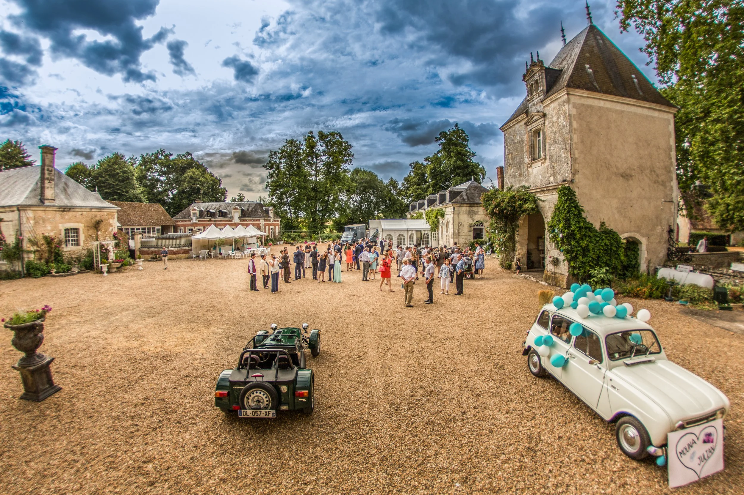 Large outdoor gathering in a courtyard surrounded by historic buildings, with groups of people socializing, some dressed formally, and decorated vintage cars.
