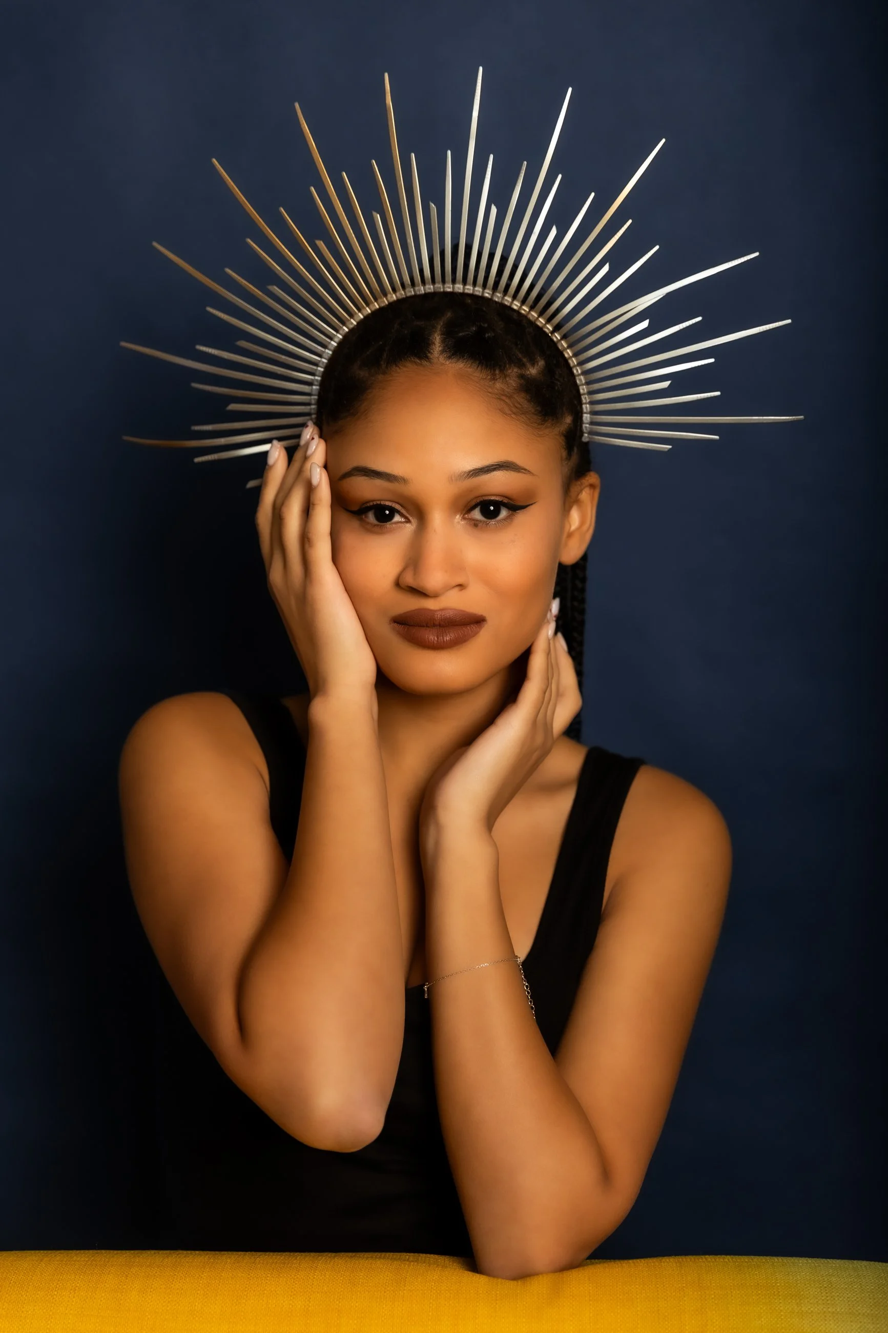 A woman with dark hair styled in braids, wearing makeup with winged eyeliner and brown lipstick, resting her hand on her face. She is wearing a black top and a thin bracelet on her wrist, with a decorative metallic halo headpiece around her head, aga