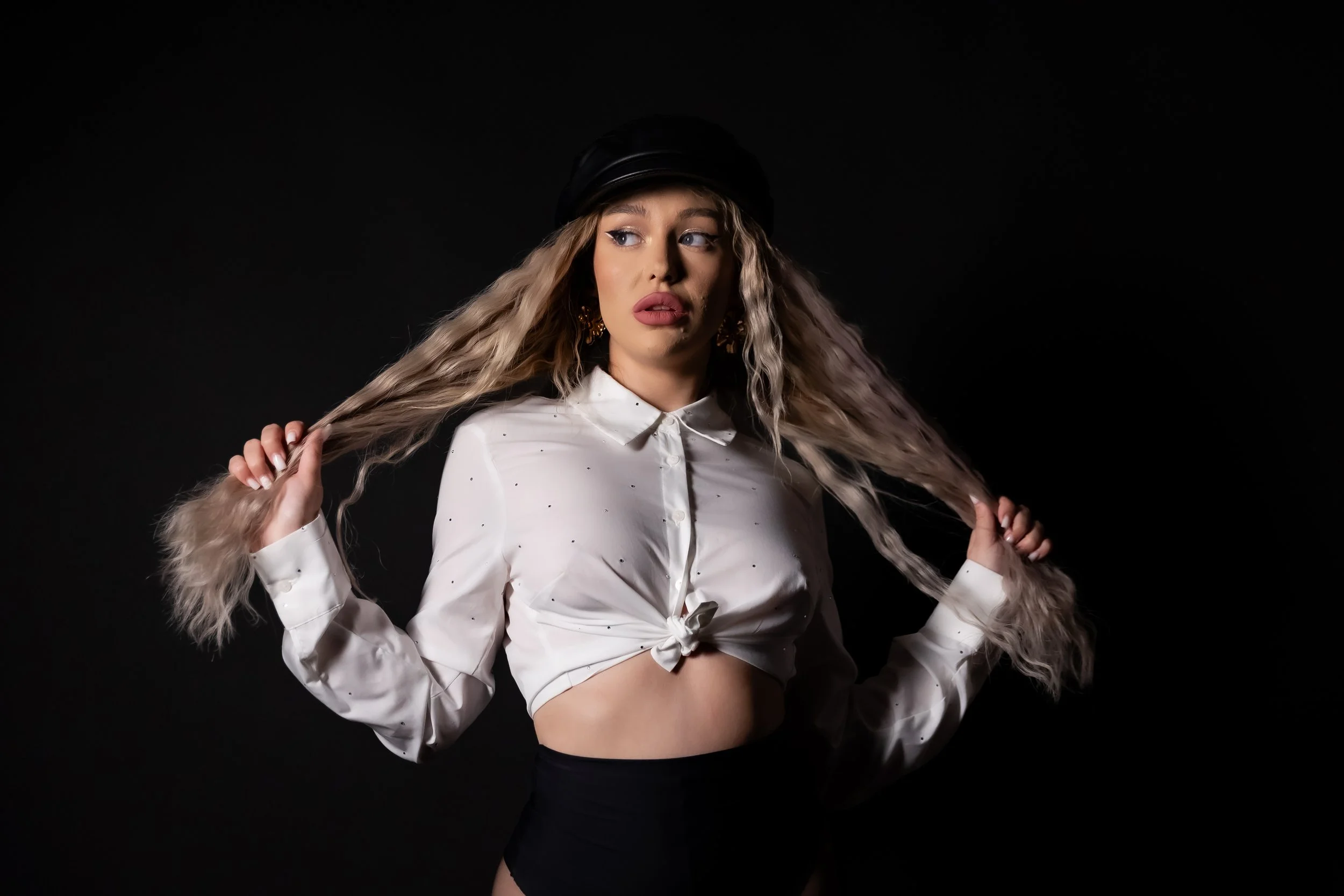 A woman with long wavy blonde hair in a tied white blouse, black high-waisted shorts, and gold earrings, holding strands of her hair against a dark background.