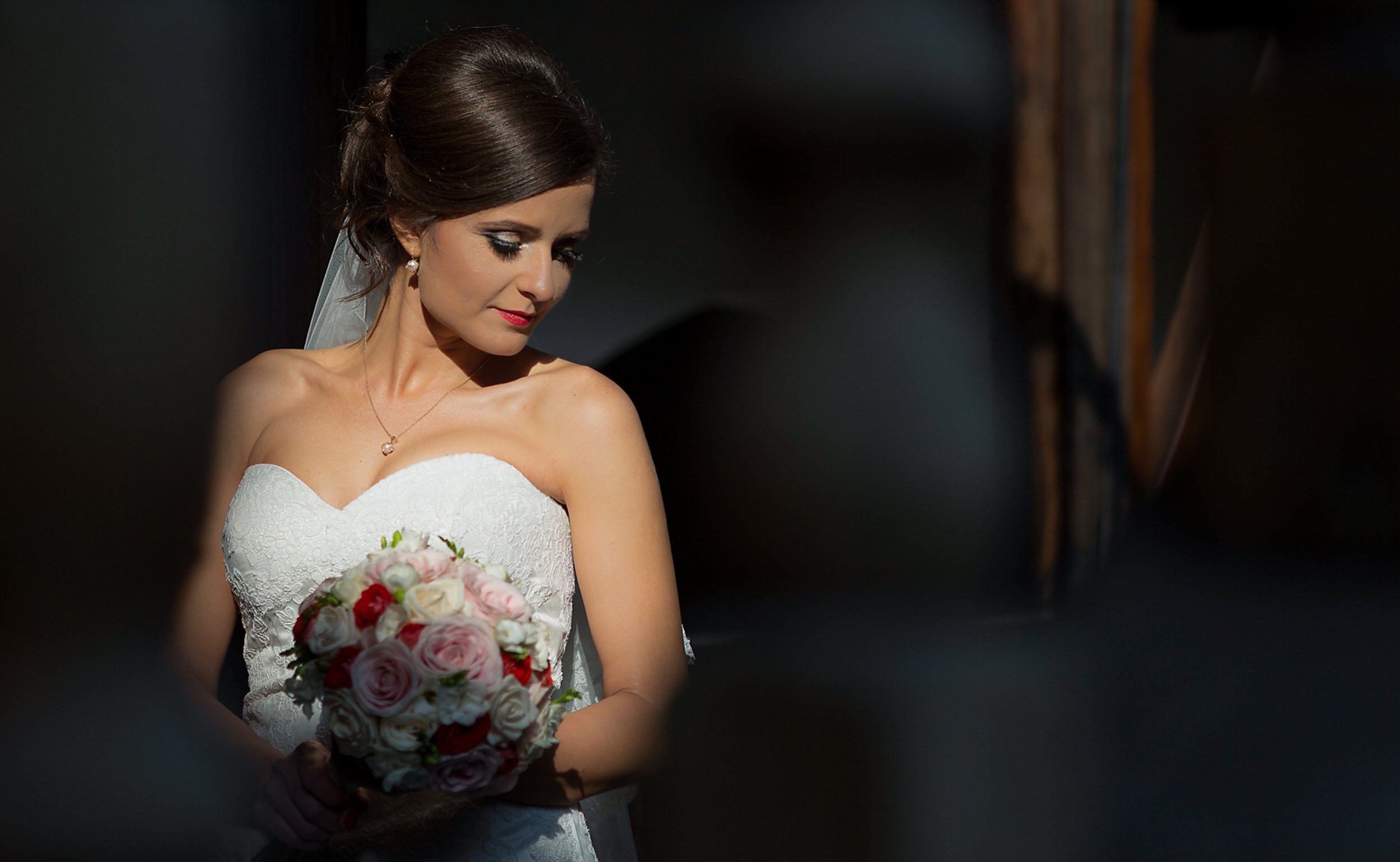 A bride with dark hair styled in an updo, wearing a strapless white lace wedding gown, holding a bouquet of pink and white roses, looking down.