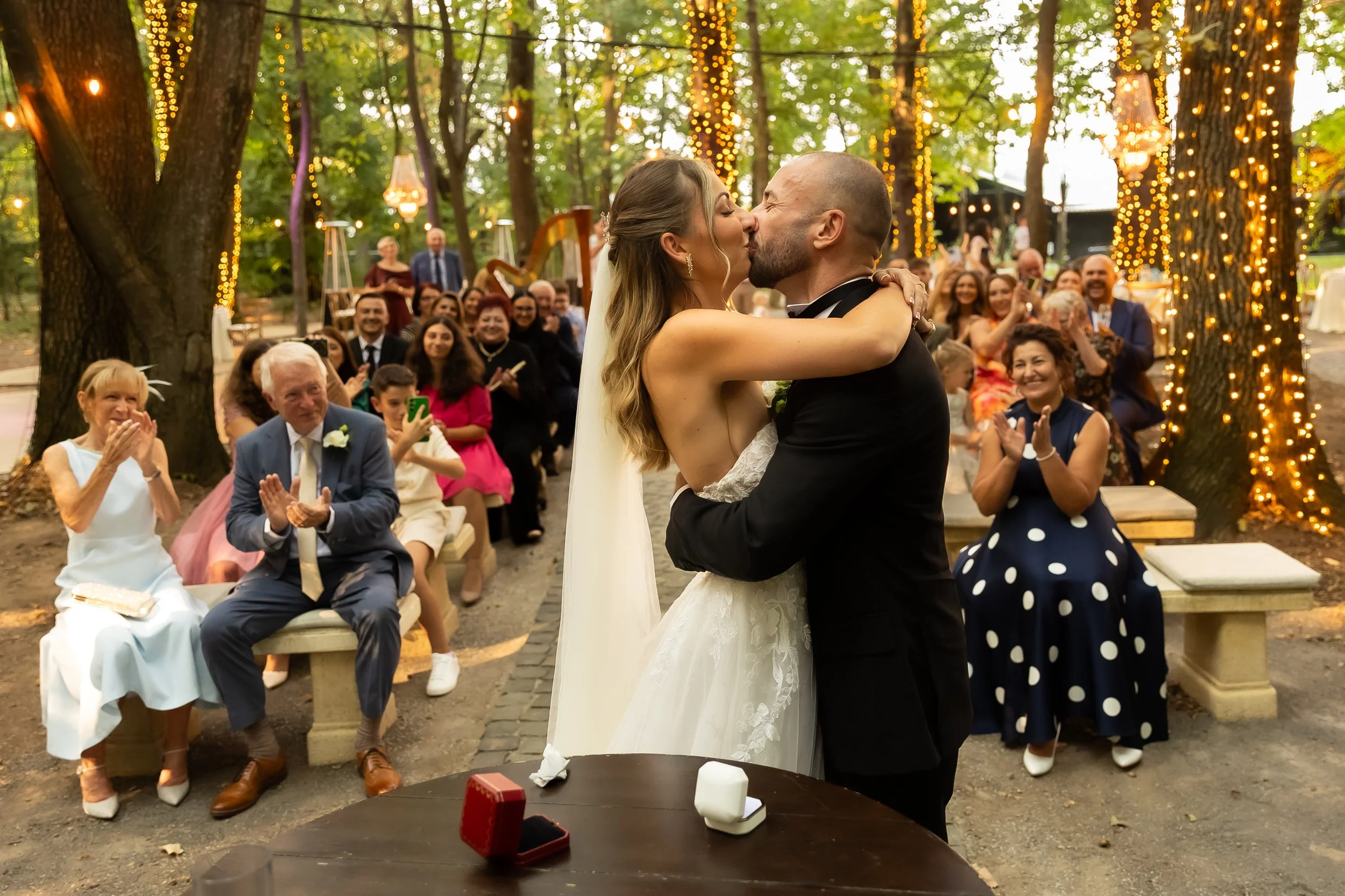A bride and groom kiss during their outdoor wedding ceremony, with guests seated on benches and surrounded by trees decorated with string lights.