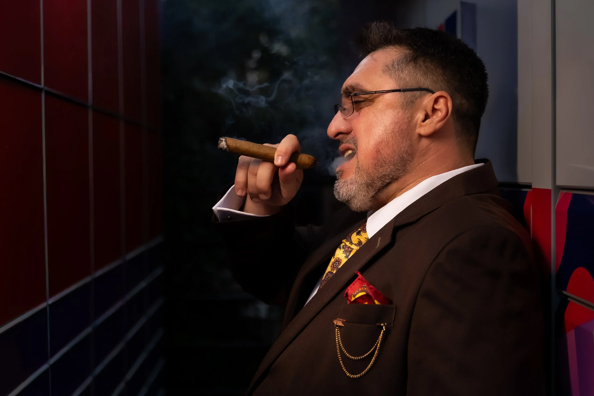 A man with glasses and a beard, smiling, holding a cigar in his right hand near his mouth, wearing a dark suit with a patterned shirt and a red pocket square, standing outdoors next to a wall with dark red and gray tiles.