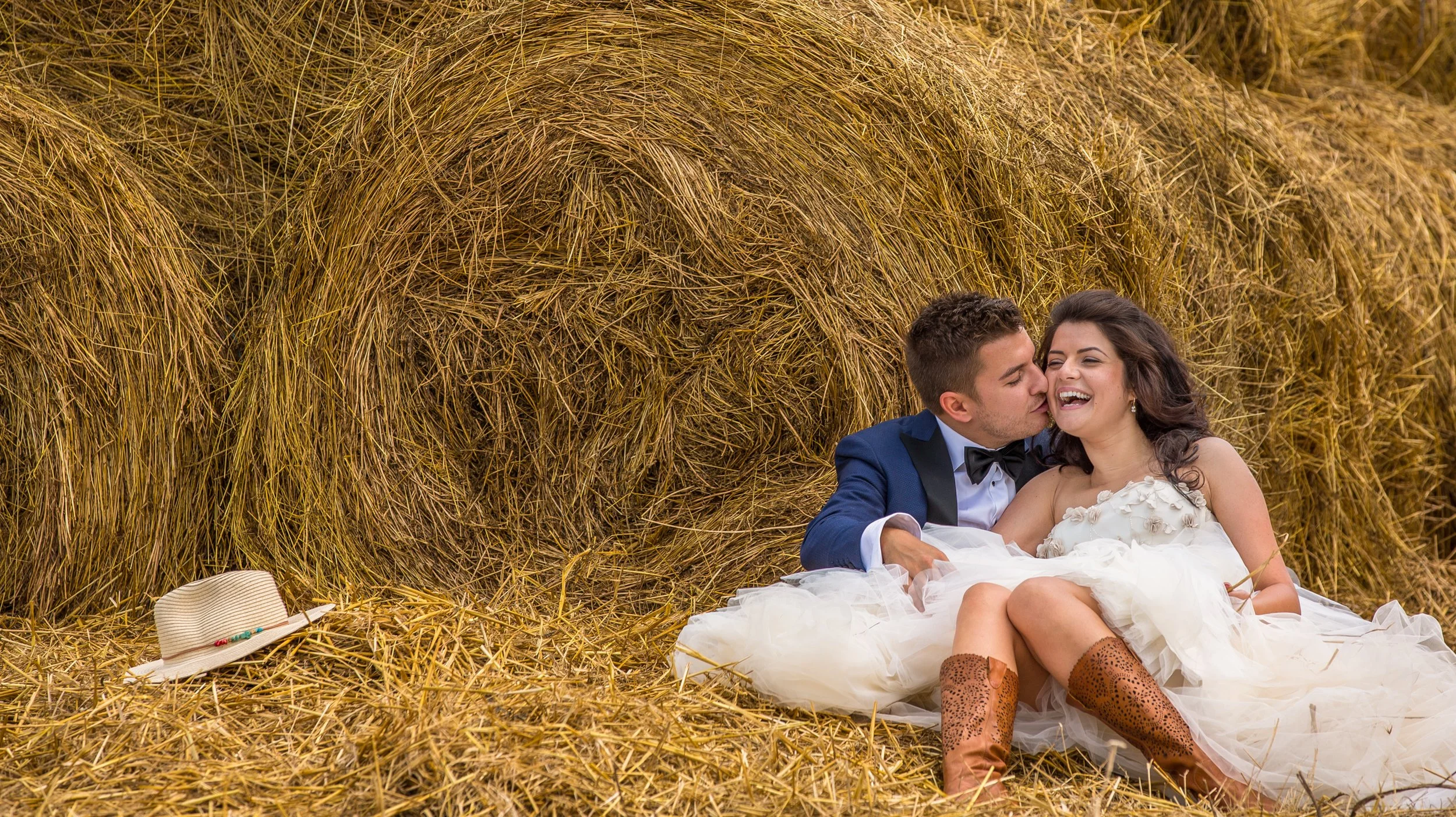 A couple in wedding attire sitting on the ground in front of hay bales, with a hat and jewelry nearby, smiling and sharing an intimate moment.