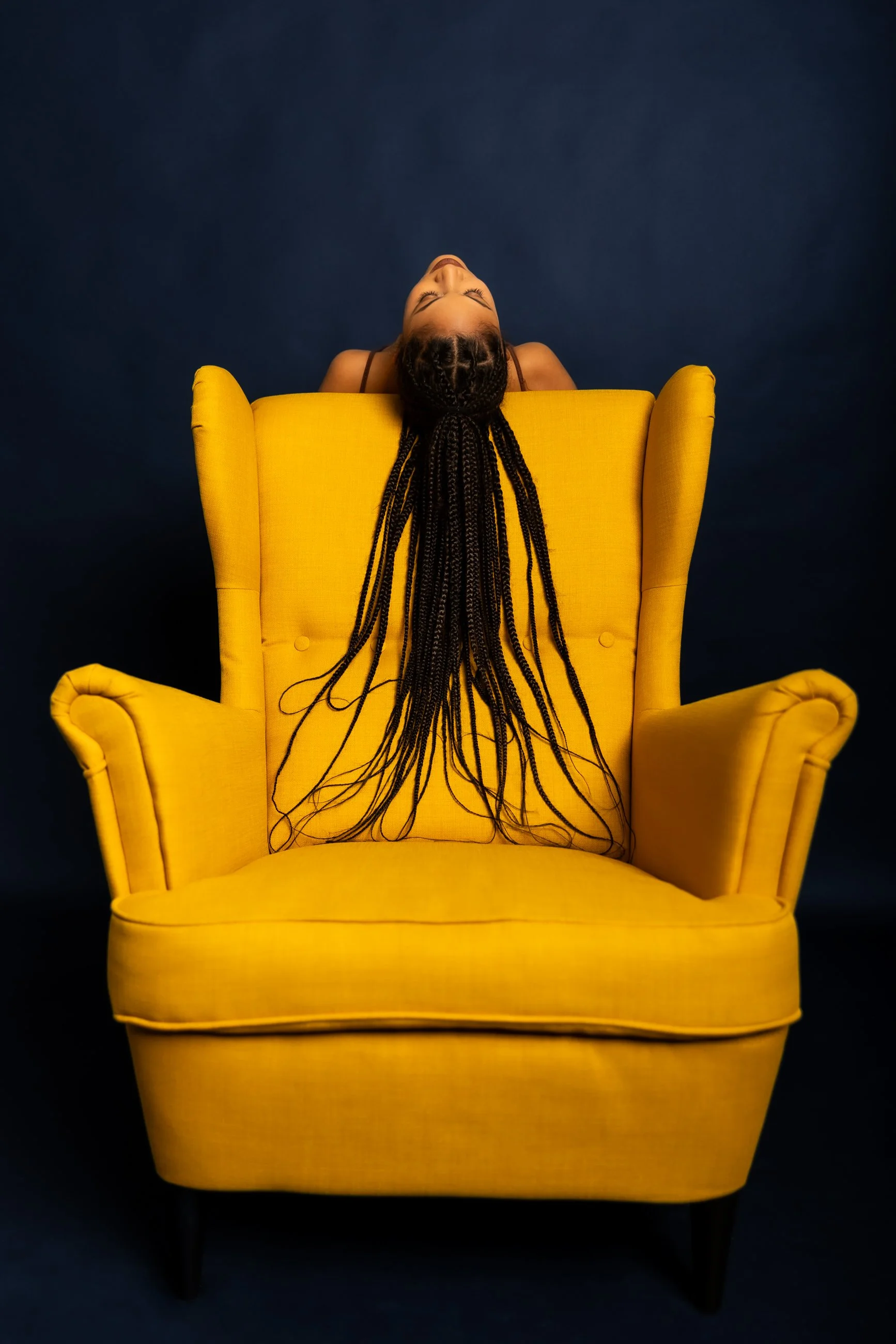 A person with braided hair sitting with their head tilted back in a yellow armchair against a dark background.