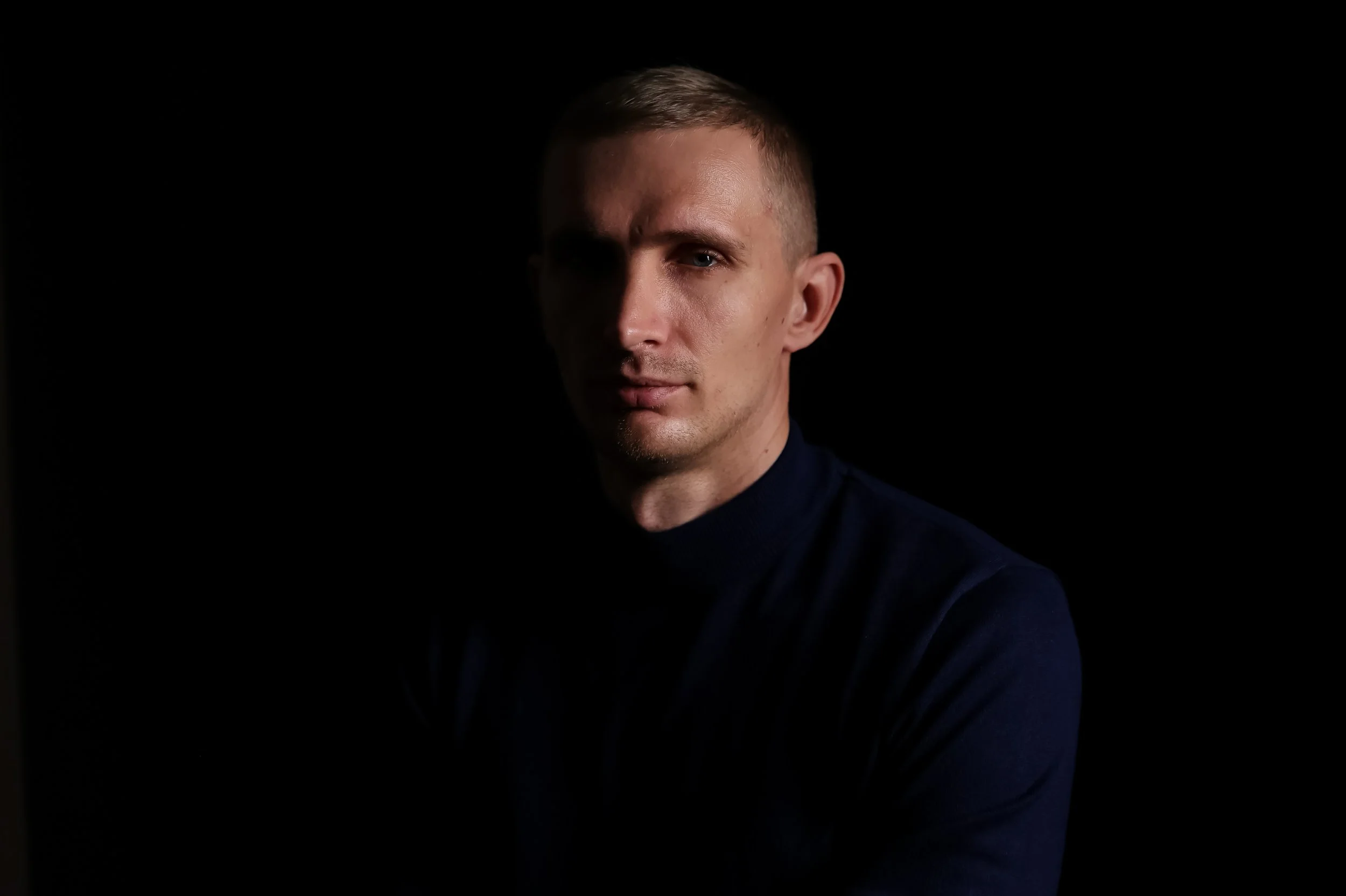 A portrait of a young man with short hair, wearing a dark turtleneck sweater, against a black background.