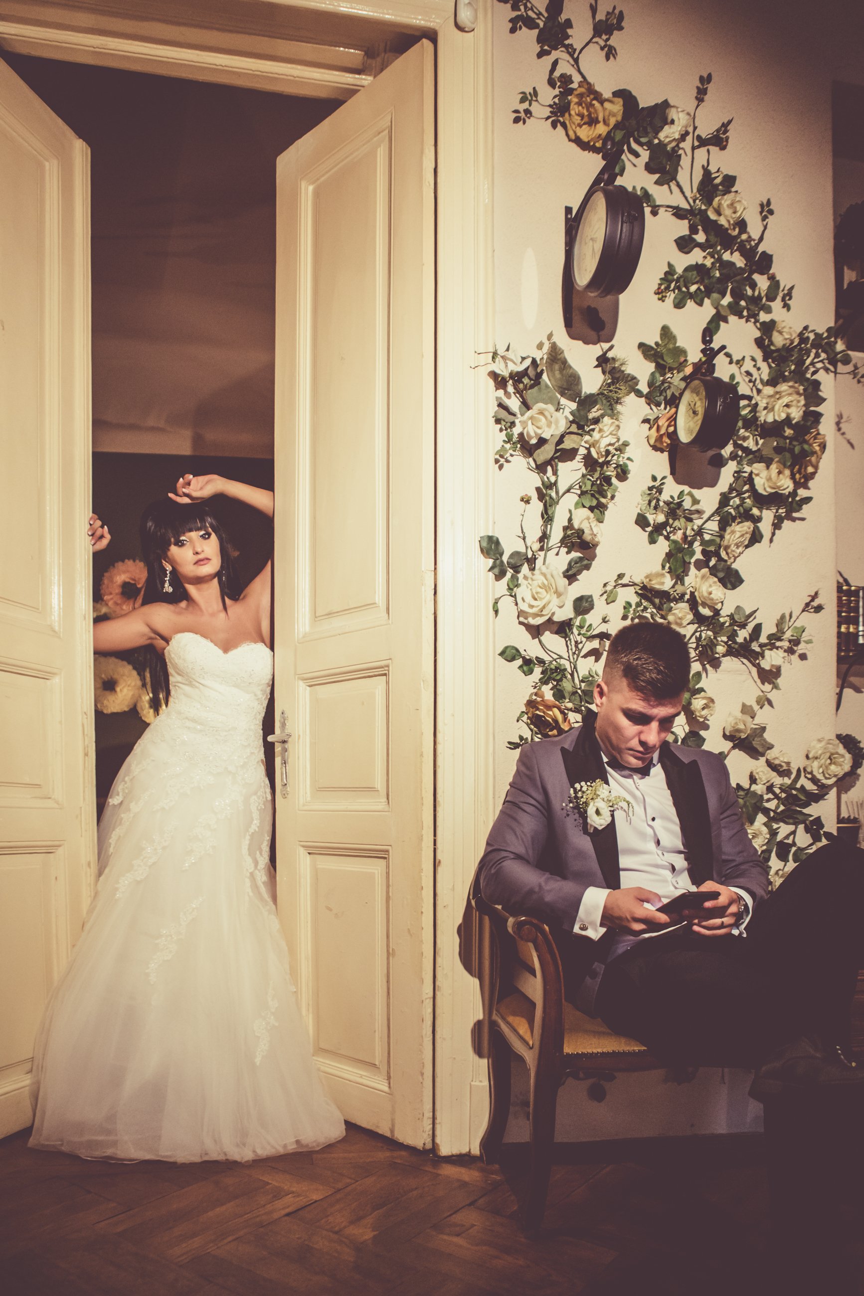 A bride with dark hair and makeup in a white wedding dress standing in a doorway with open doors. A groom in a gray tuxedo is sitting on a chair, looking at his phone in a decorated room with floral and clock wall decorations.