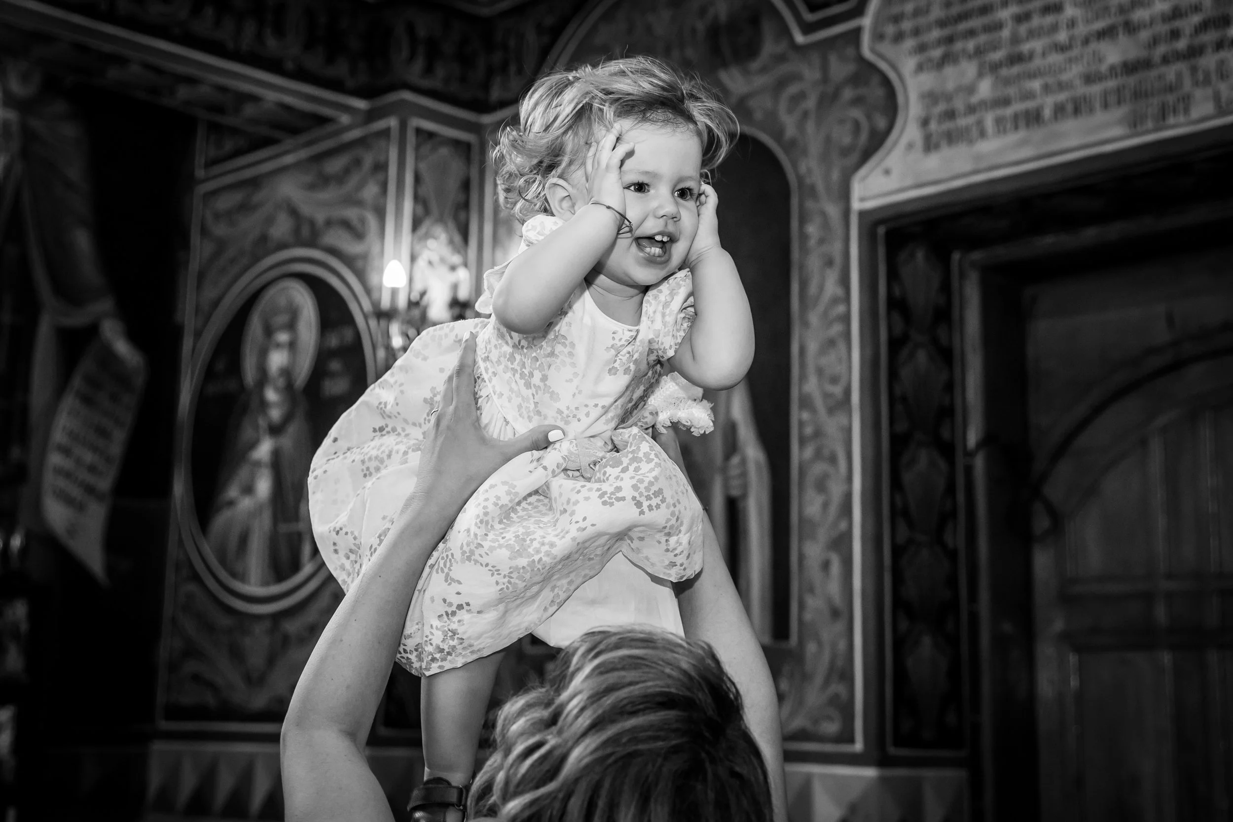 A young girl with curly hair and a floral dress is held up by an adult, covering her ears and showing a distressed expression. The background features ornate, patterned walls and religious icons, suggesting a religious or cultural setting.