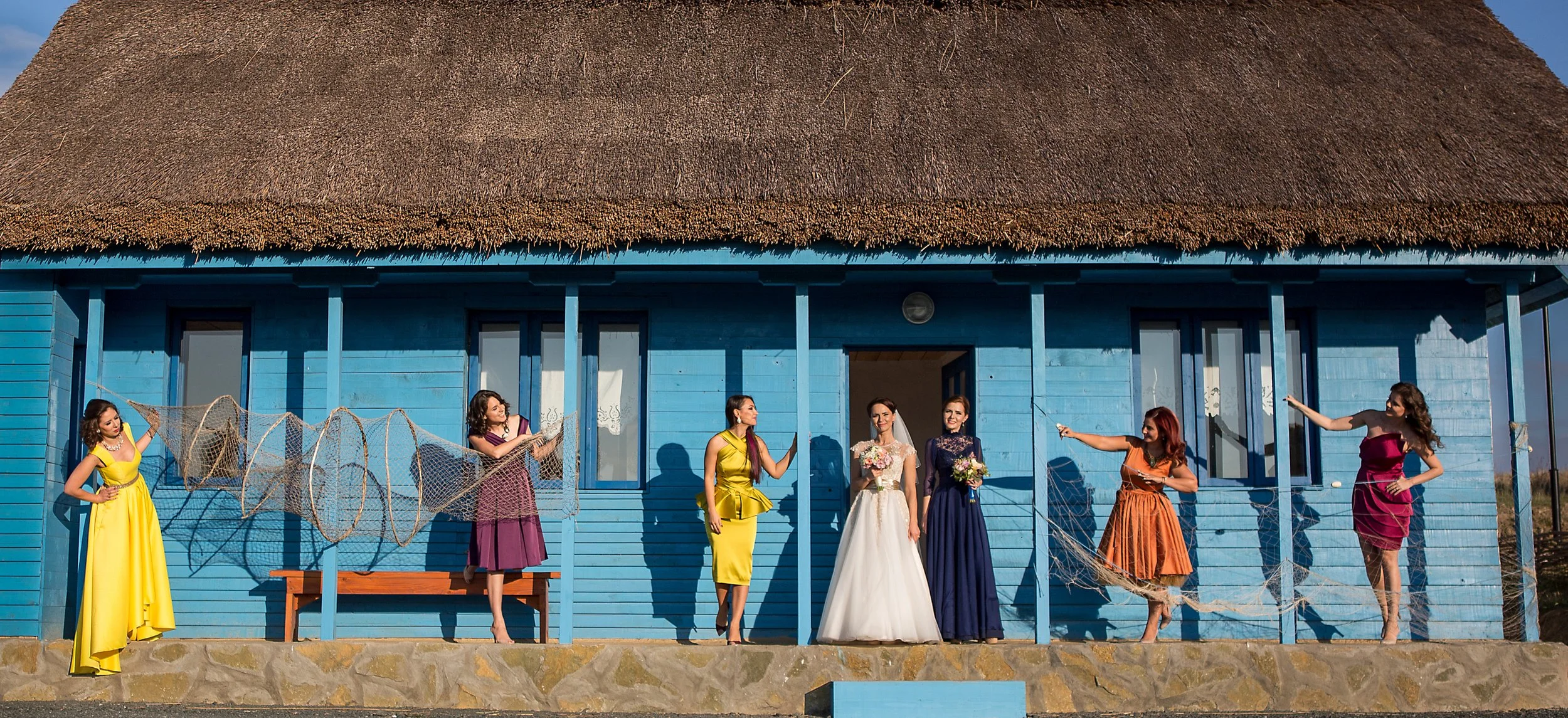 A group of women wearing colorful dresses standing in front of a blue wooden house with a thatched roof, holding fishing nets and flowers, during daytime.