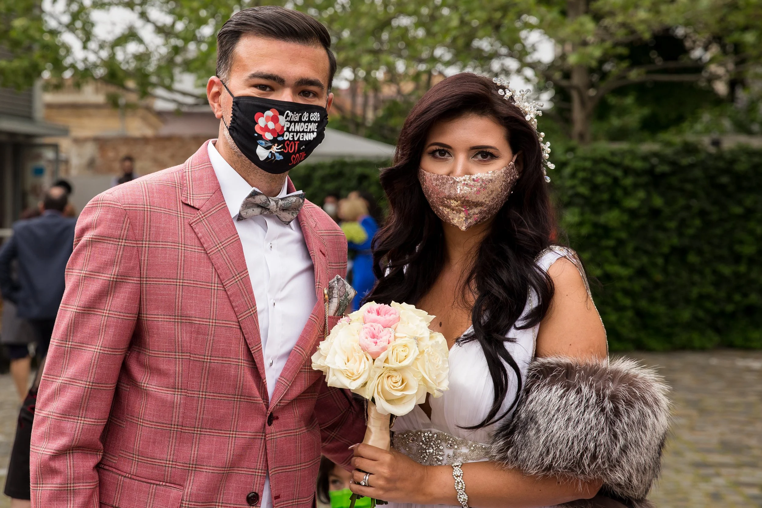 A man and woman dressed in wedding attire wearing face masks, standing outdoors surrounded by greenery, with the woman holding a bouquet of white and pink roses.