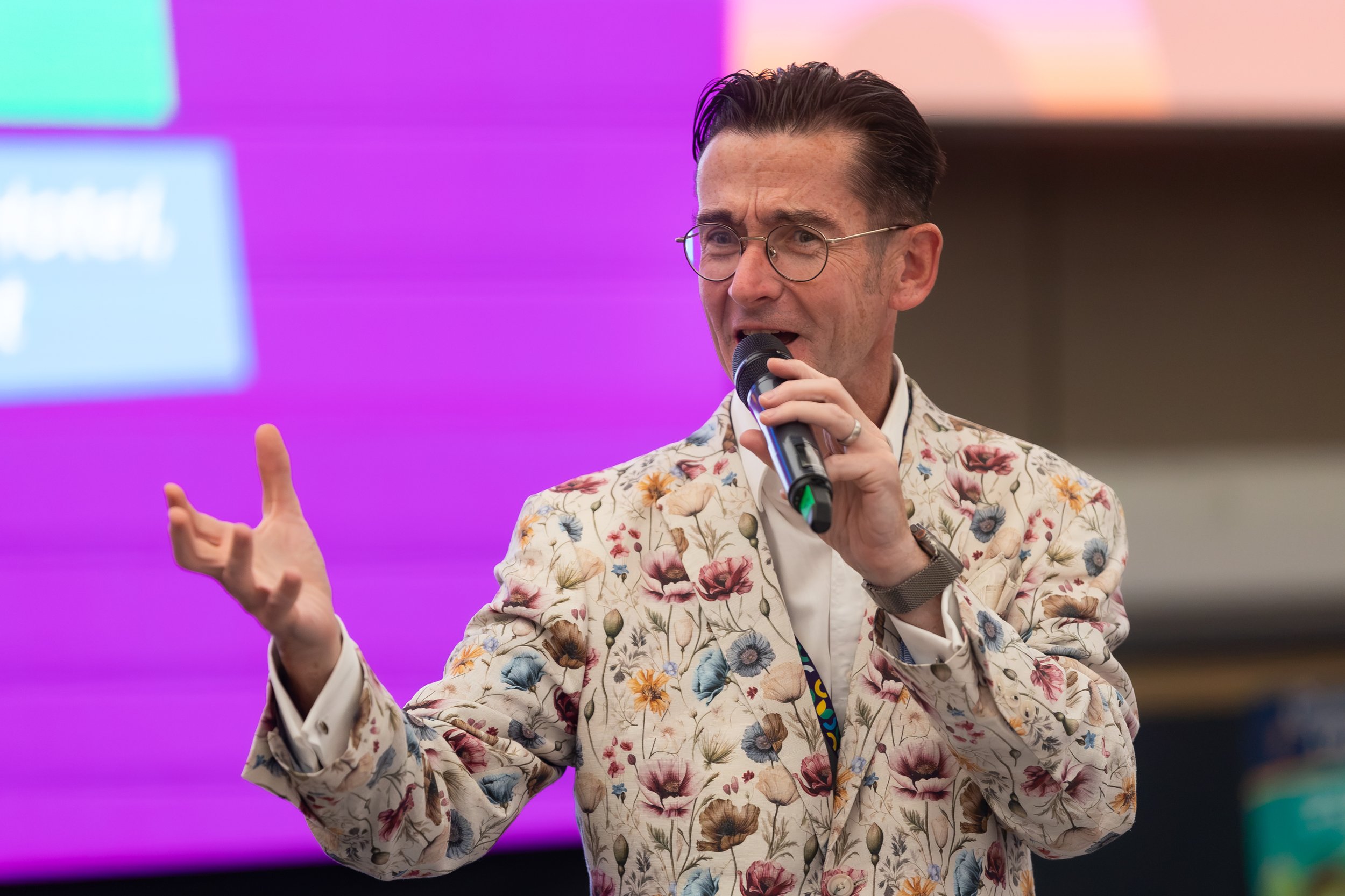 A man wearing glasses and a floral blazer holding a microphone and speaking at an event with a purple and pink background.