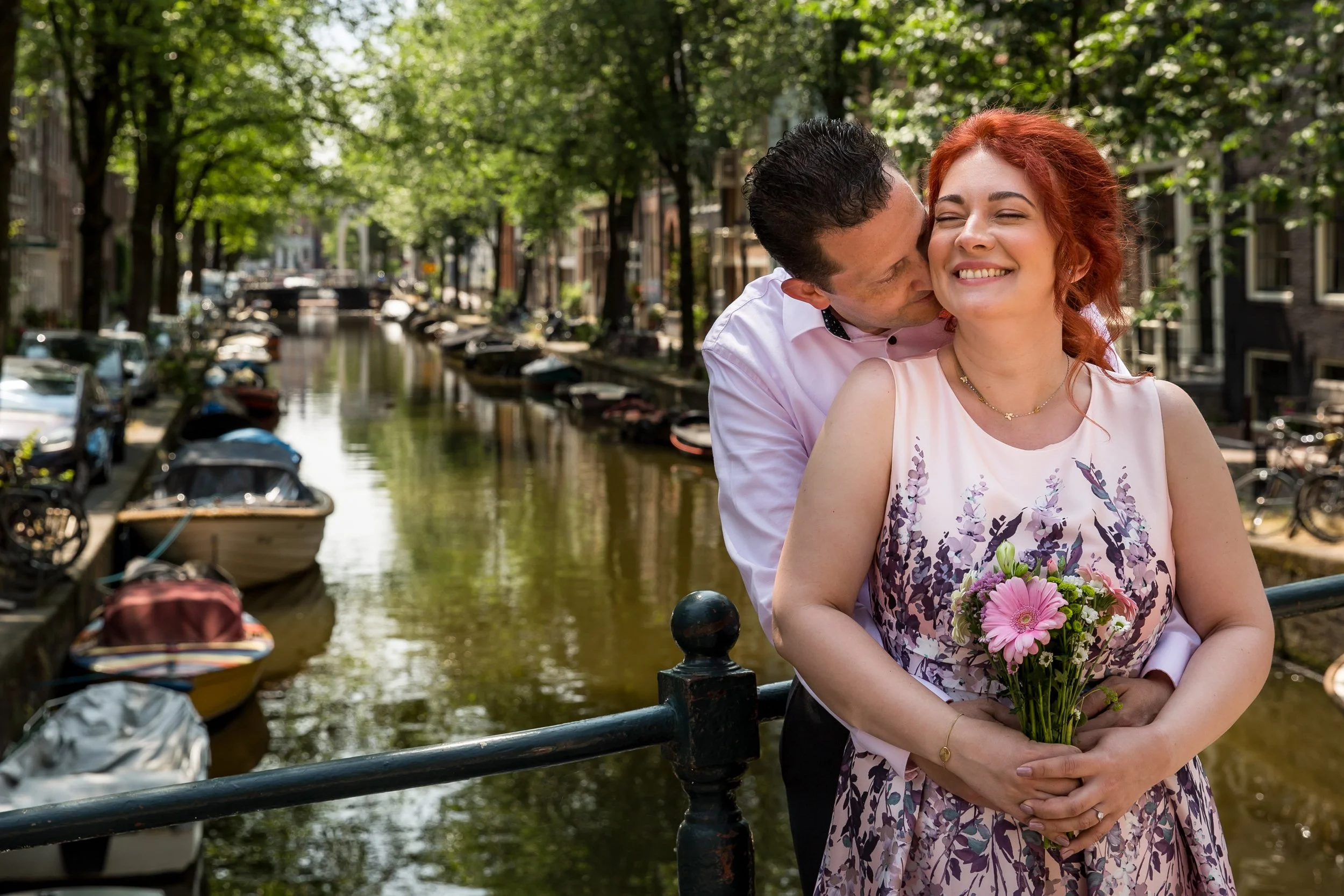 A couple standing near a canal with boats and trees, with the man kissing the woman on the cheek while she smiles holding a bouquet of flowers.