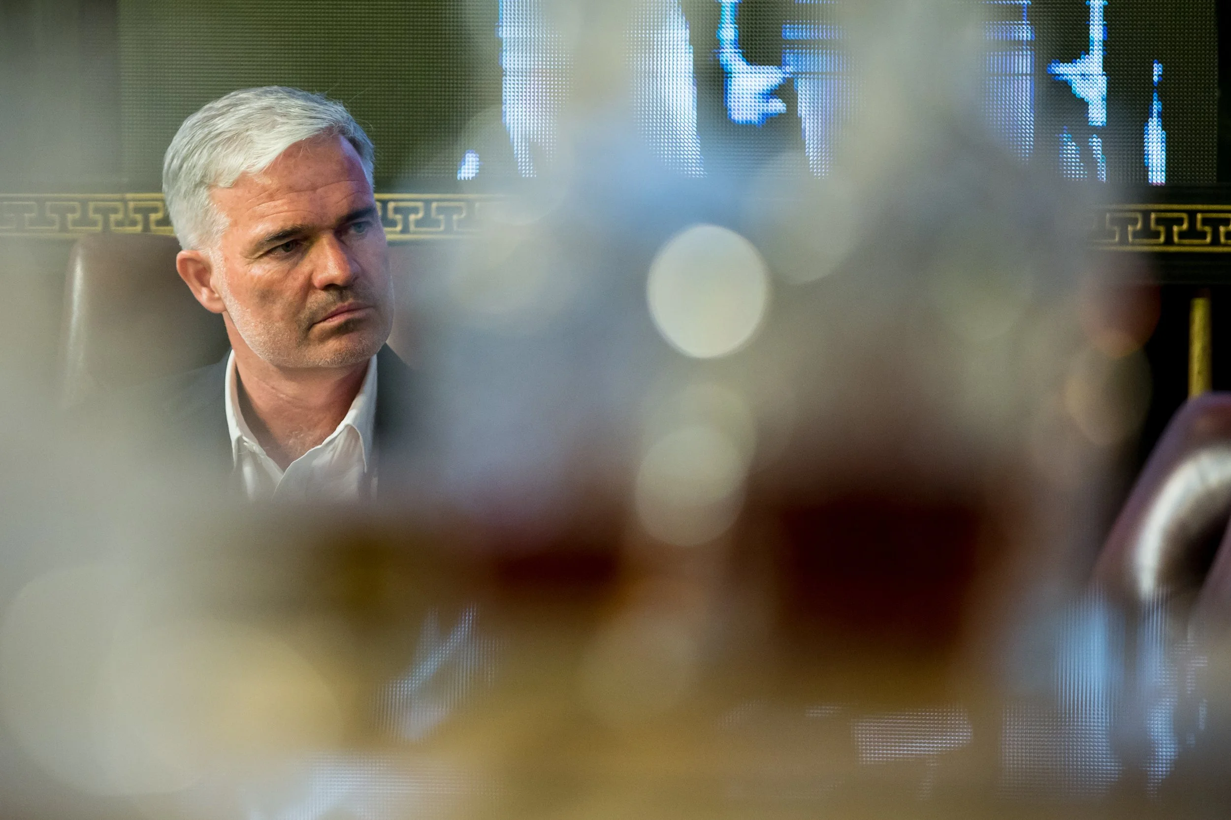 A middle-aged man with gray hair, wearing a white shirt, is seated and appears to be listening intently. The photo is taken through a semi-transparent surface with bokeh light effects and out-of-focus elements in the foreground.