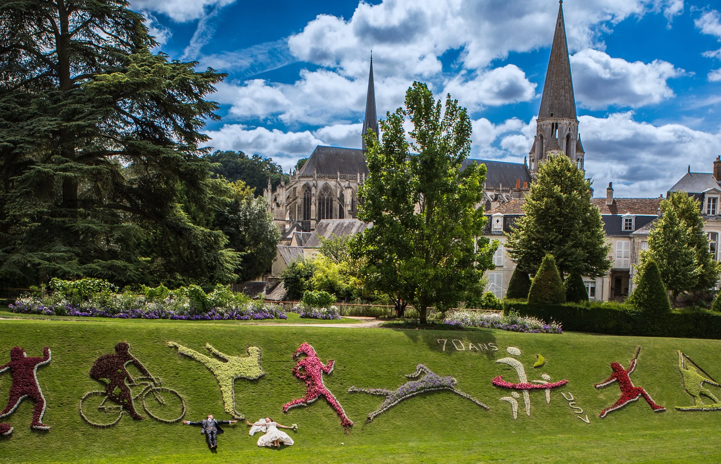 Colorful flower garden with the words '70 ans' and cats and dogs made of flowers, with a large Gothic-style cathedral with twin spires in the background and a bright blue sky with scattered clouds.