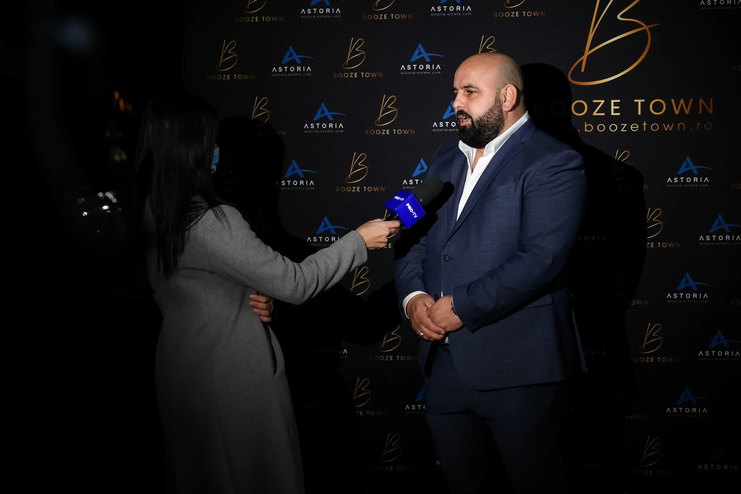 A man with a beard and bald head wearing a blue suit is being interviewed by a woman with long dark hair, who is holding a microphone labeled "PRO TV." They are standing in front of a black backdrop with logos for "Booze Town," "Astoria," and "astori