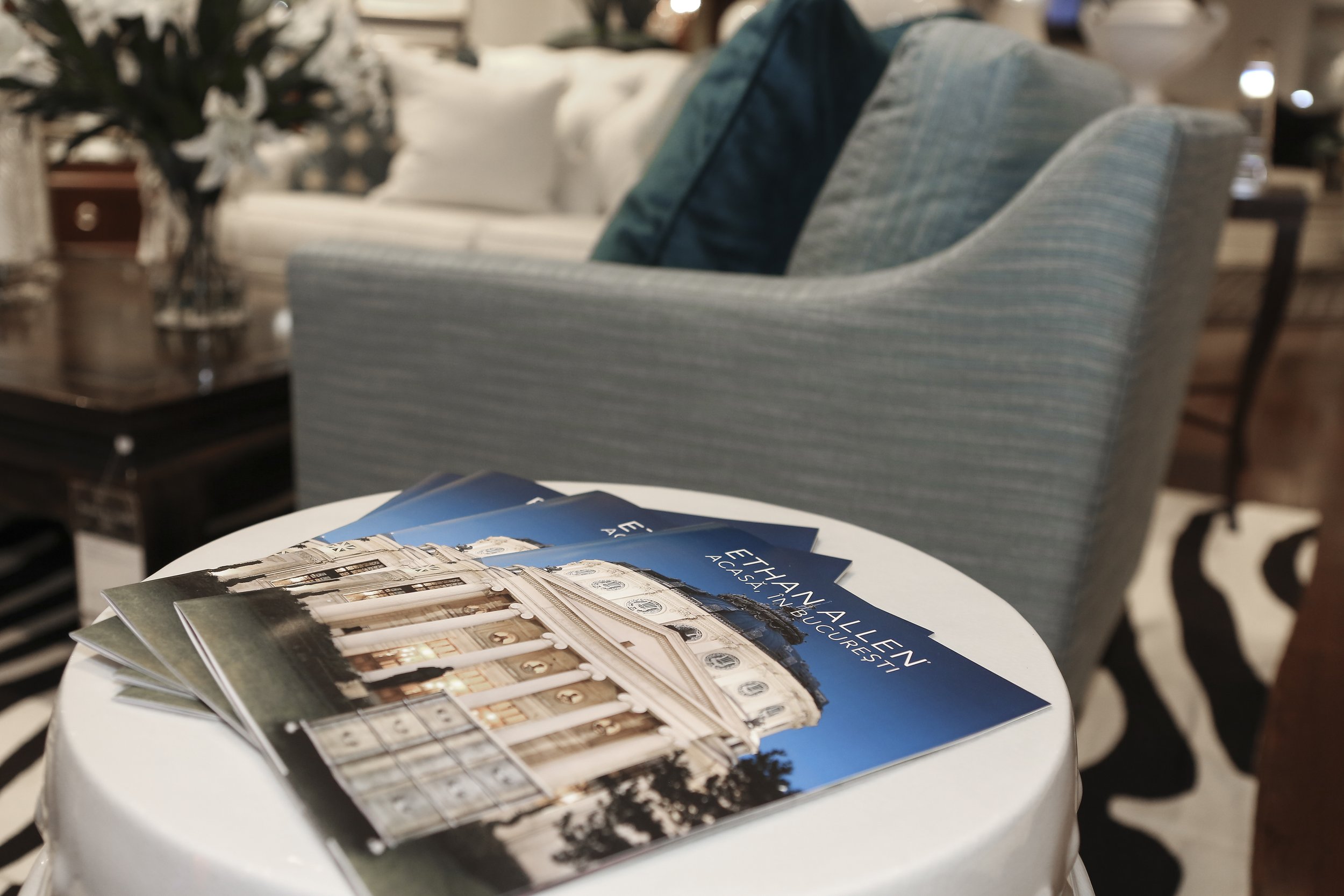 Travel brochures on a small round table in a retail store, with a background of a blue striped armchair and blurred decorative items.