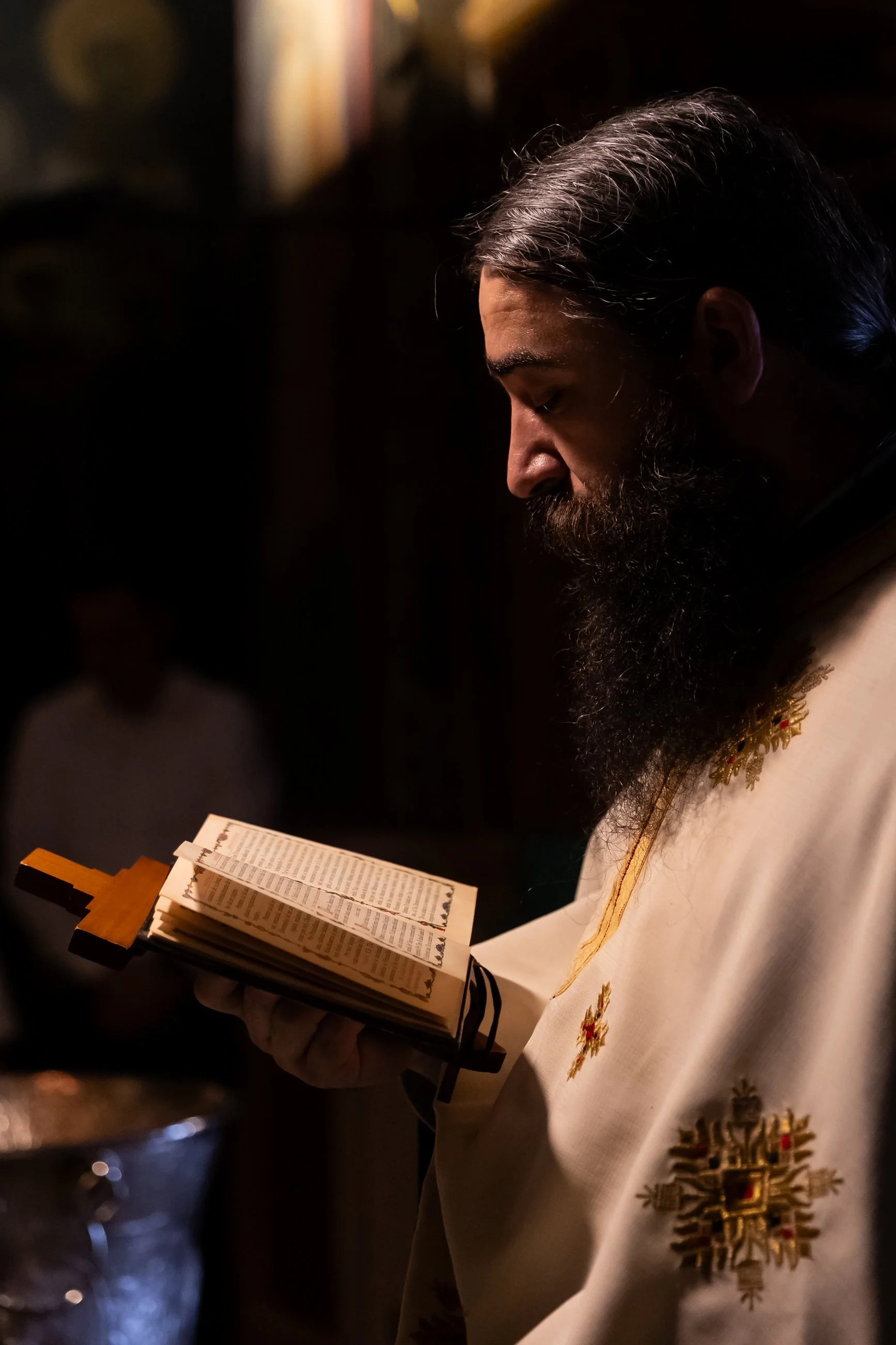 A man with long dark hair and a beard reading from a book, possibly a religious text or prayer book, adorned with a gold chain, in a dimly lit setting.