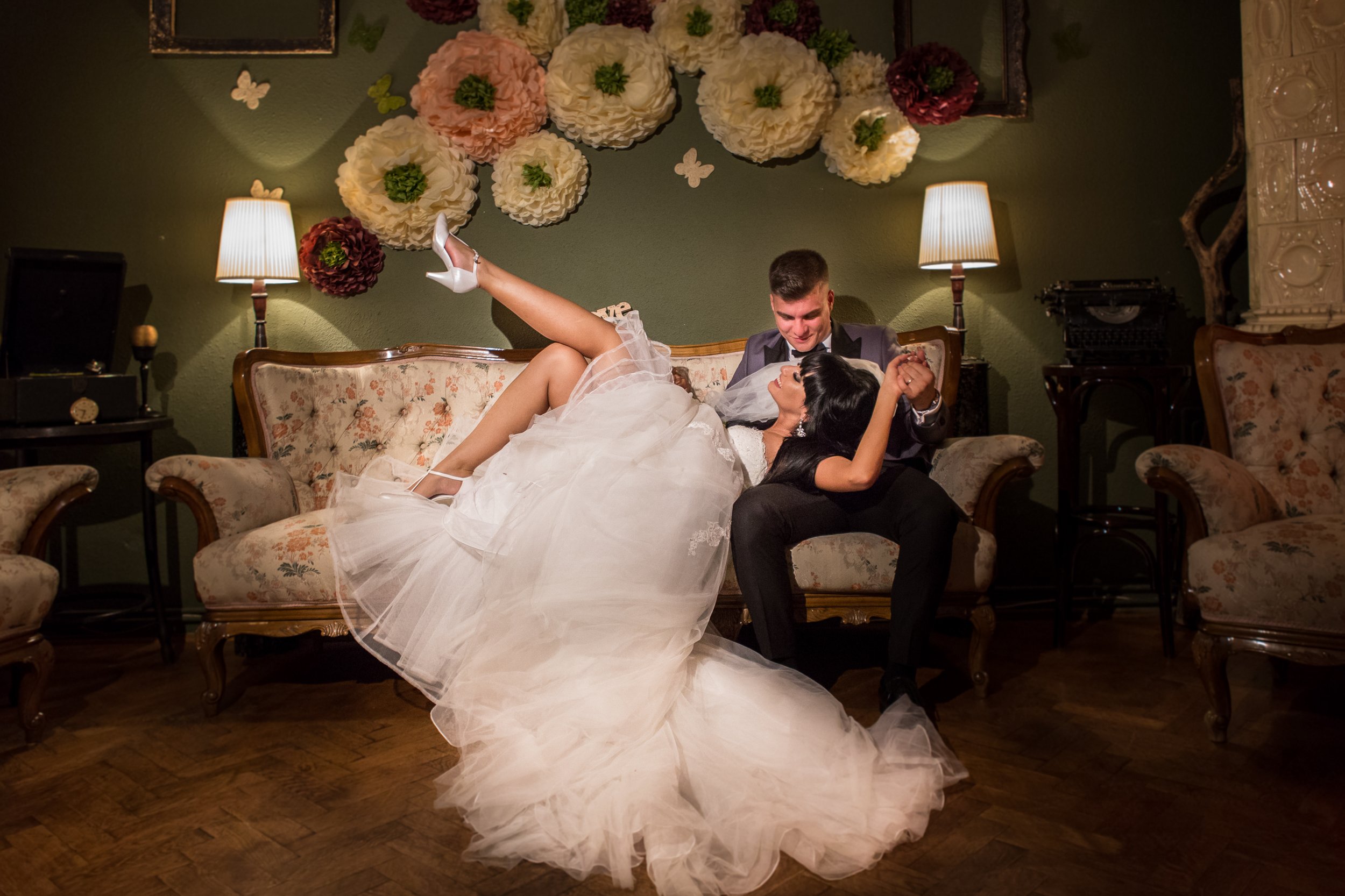 A bride and groom dancing in a living room decorated with large paper flowers and butterflies on the wall. The bride is wearing a white wedding dress and white heels, while the groom is in a dark suit and bow tie. The bride is lying on the sofa with 