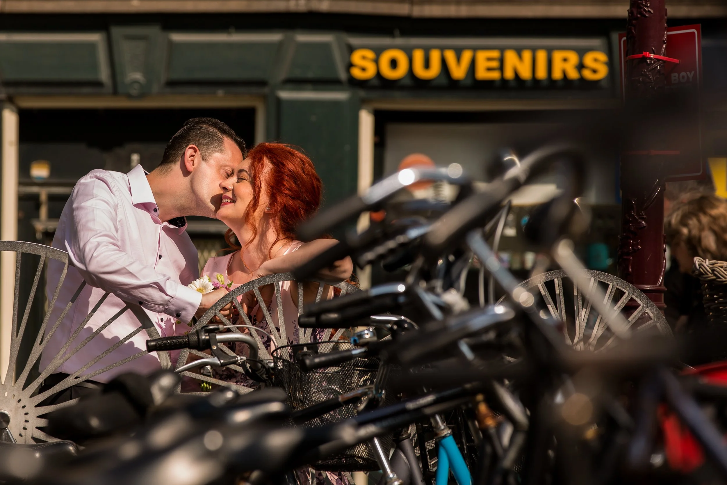 A couple sitting closely at an outdoor café, smiling and leaning in for a kiss, with a "SOUVENIRS" shop sign in the background and bicycles in the foreground.