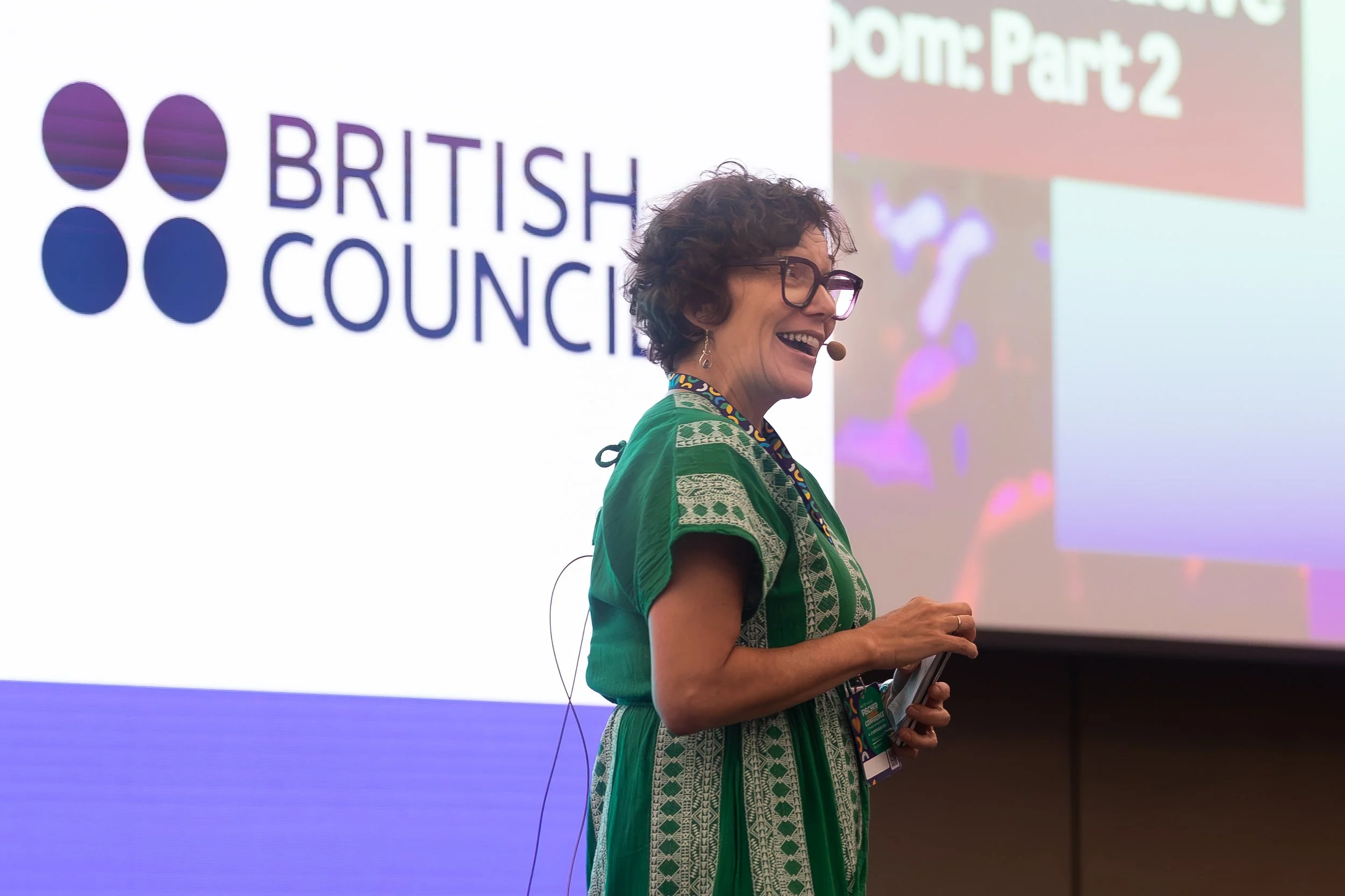 A woman with short curly hair and glasses speaking at a conference, standing in front of a large screen displaying the British O Council logo and a presentation slide.