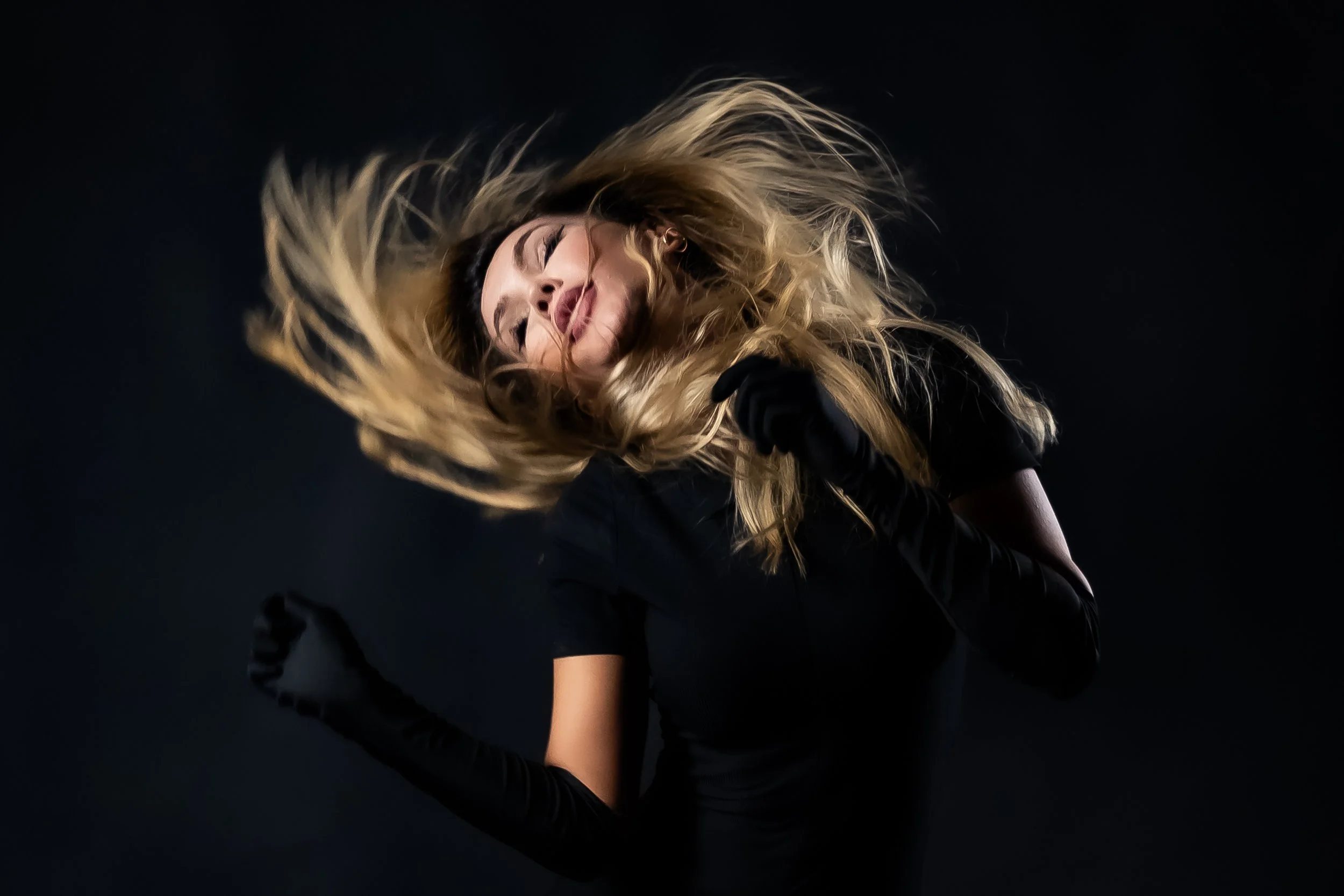 A woman with long blonde hair, wearing black gloves and black clothing, appears to be singing or performing against a dark background, with her hair flowing around her face.