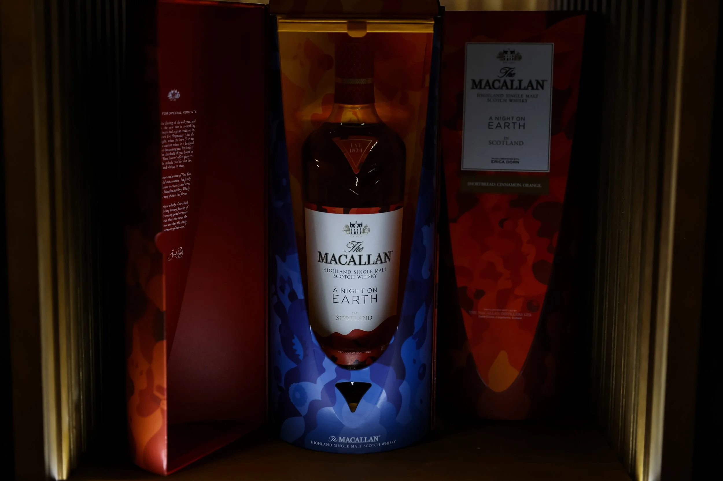 A bottle of The Macallan Highland Single Malt Scotch Whisky in its original packaging, displayed on a shelf with a red and orange box behind it.