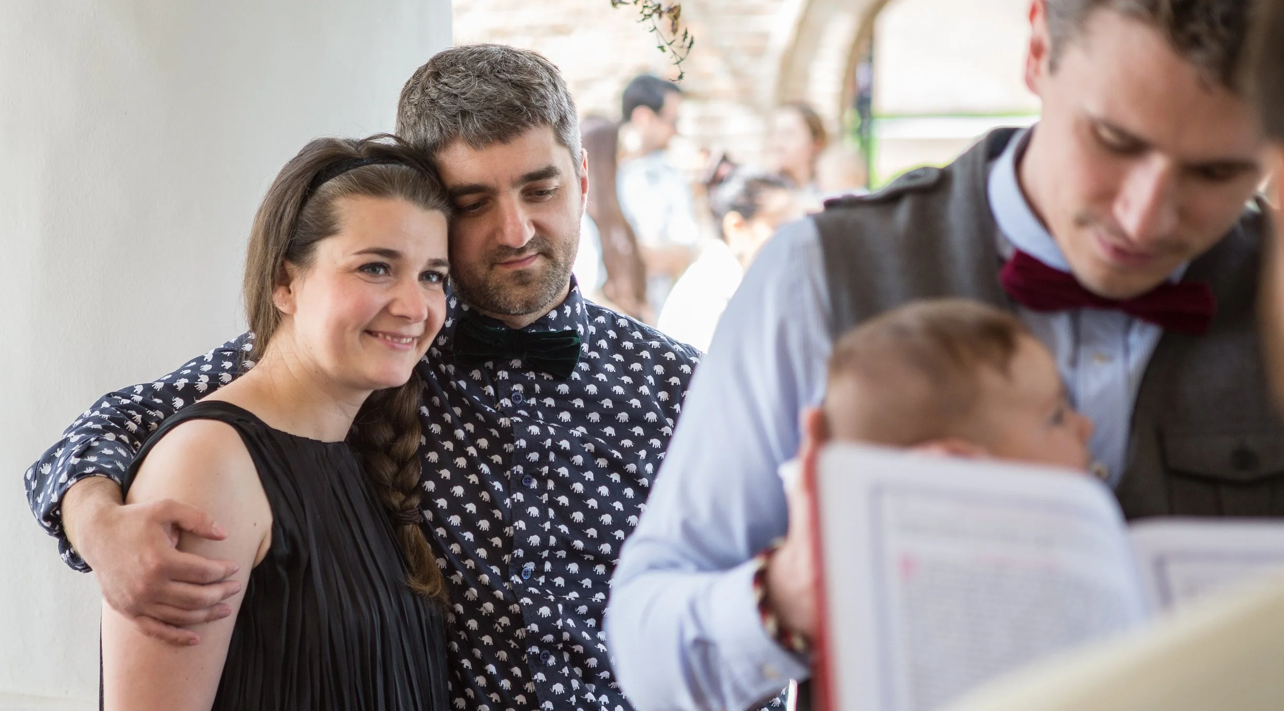 A woman and a man are happily posing together, with the woman smiling and the man resting his head on her. They are standing close to each other. In the foreground, a waiter in a uniform with a red bow tie is serving a child, who is holding a menu or