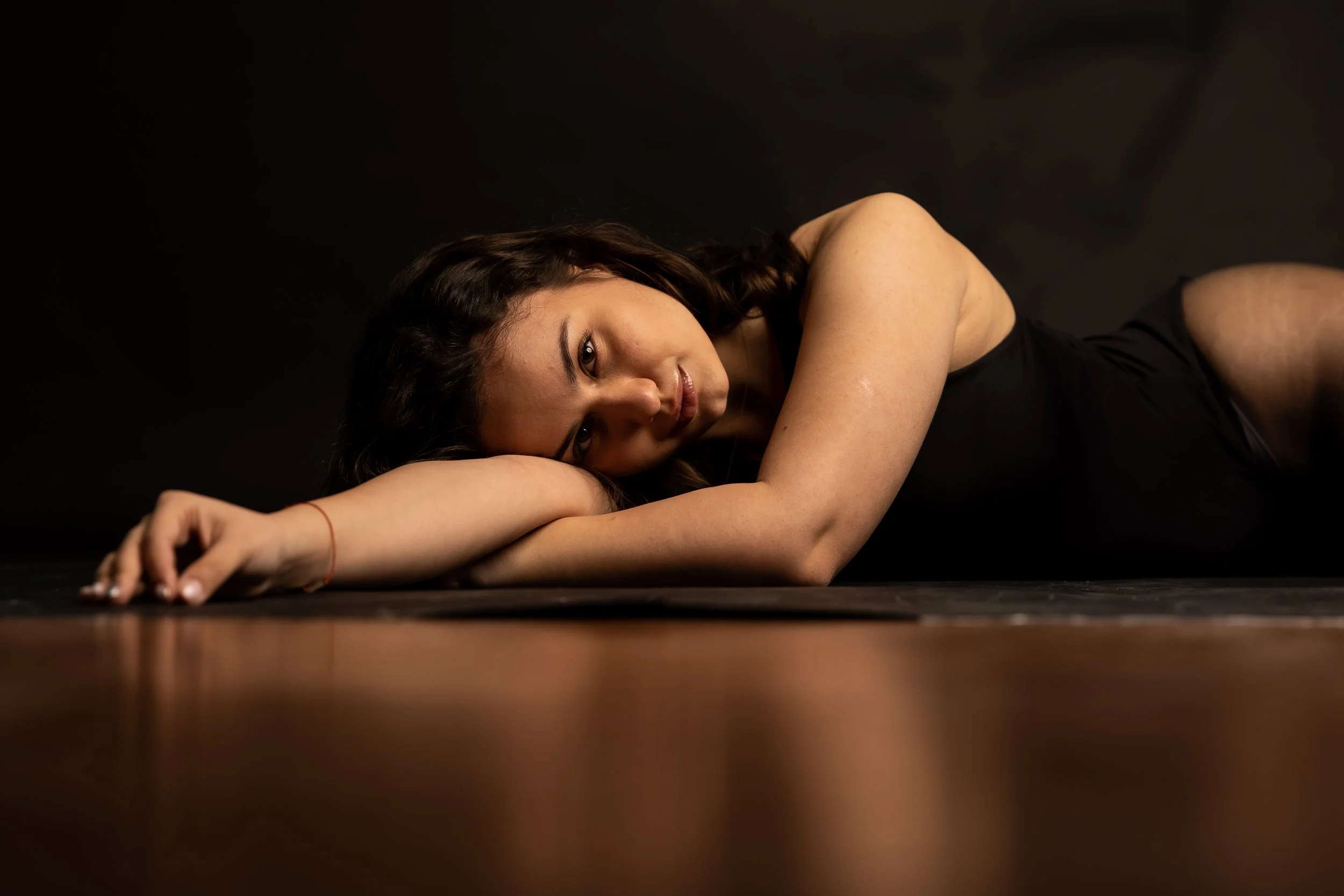 A woman with dark hair and a black top lying on the floor with a black background, resting her head on her arm, gaze directed at the camera.